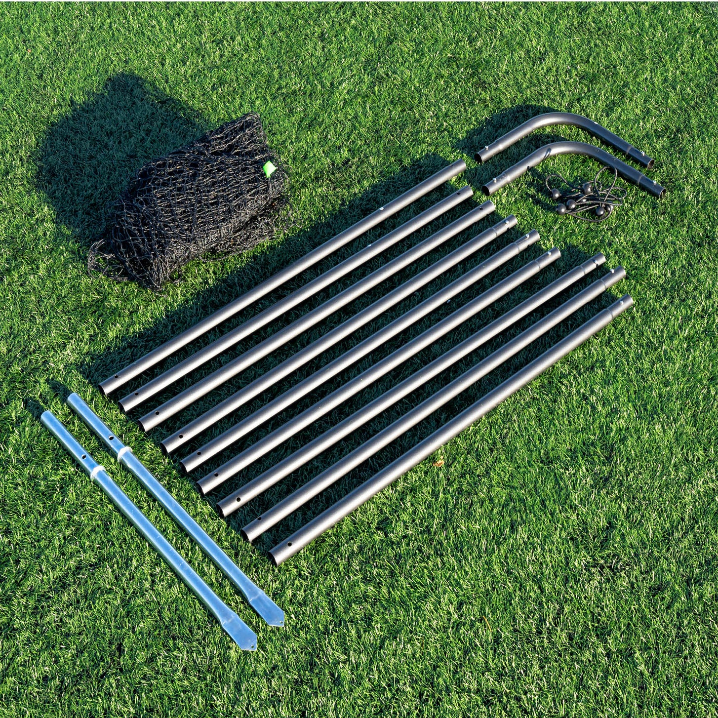 10ft Barrier Net System Freestanding parts on artificial turf: bagged black net, black frame tubes, two curved corner tubes and two silver posts.