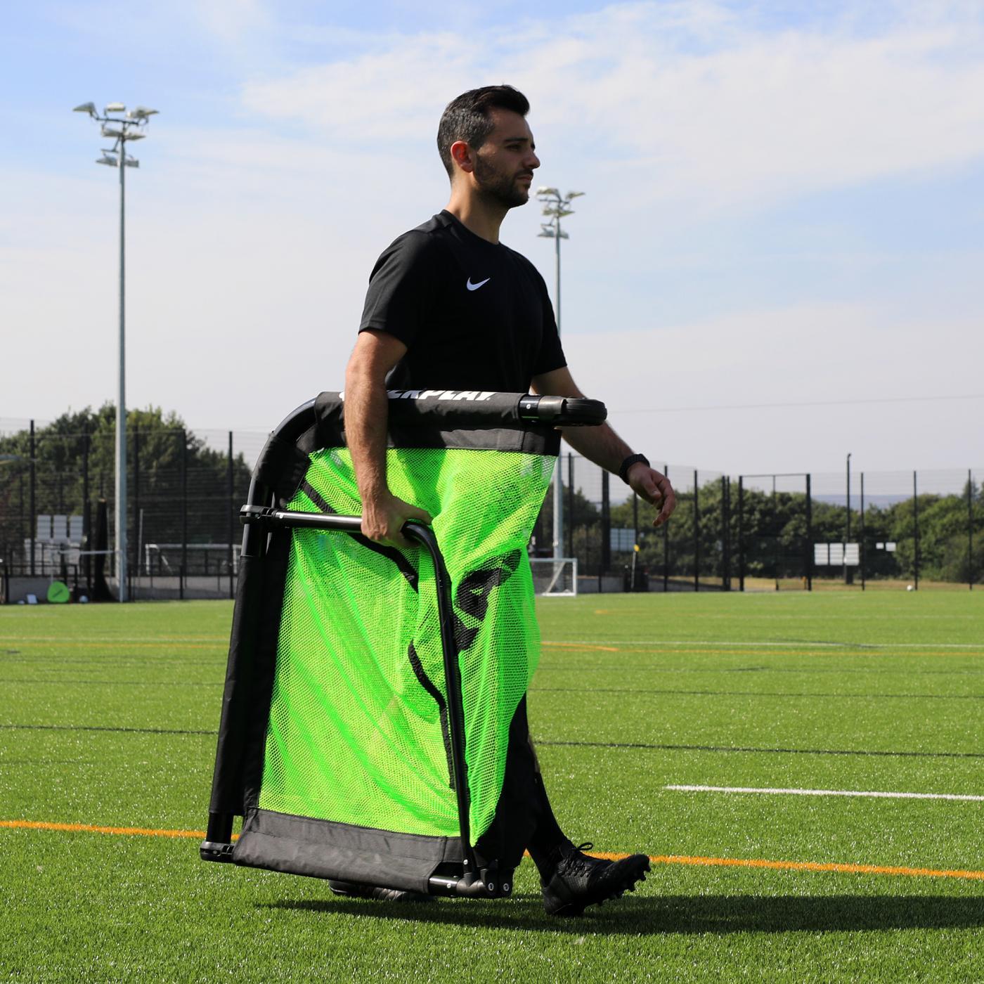 Man carrying the PRO ALU Training Soccer Goal 6x4' under one arm across an artificial field.
