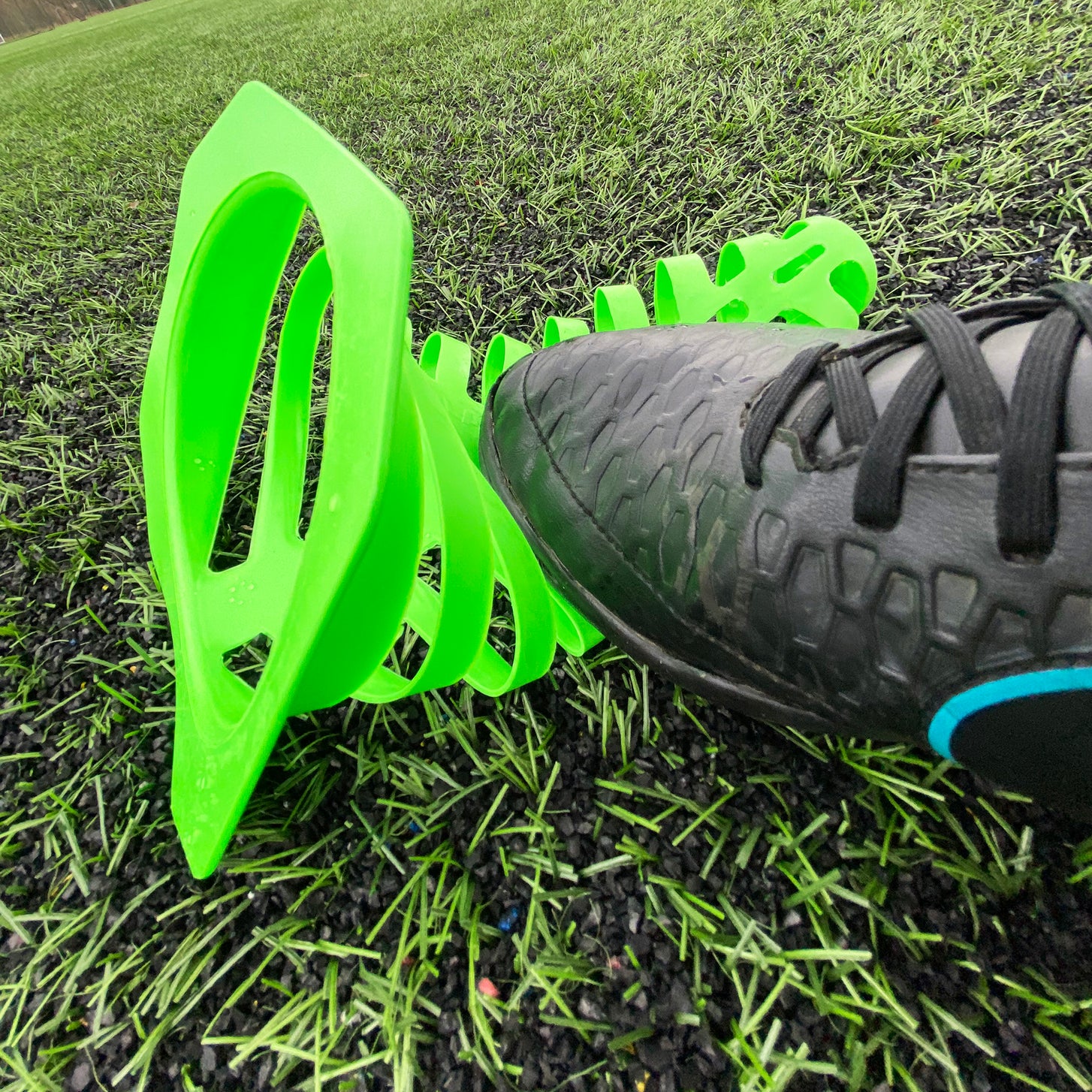Green 9" Tall Collapsible Cone squashed flat under a black cleat on artificial turf.
