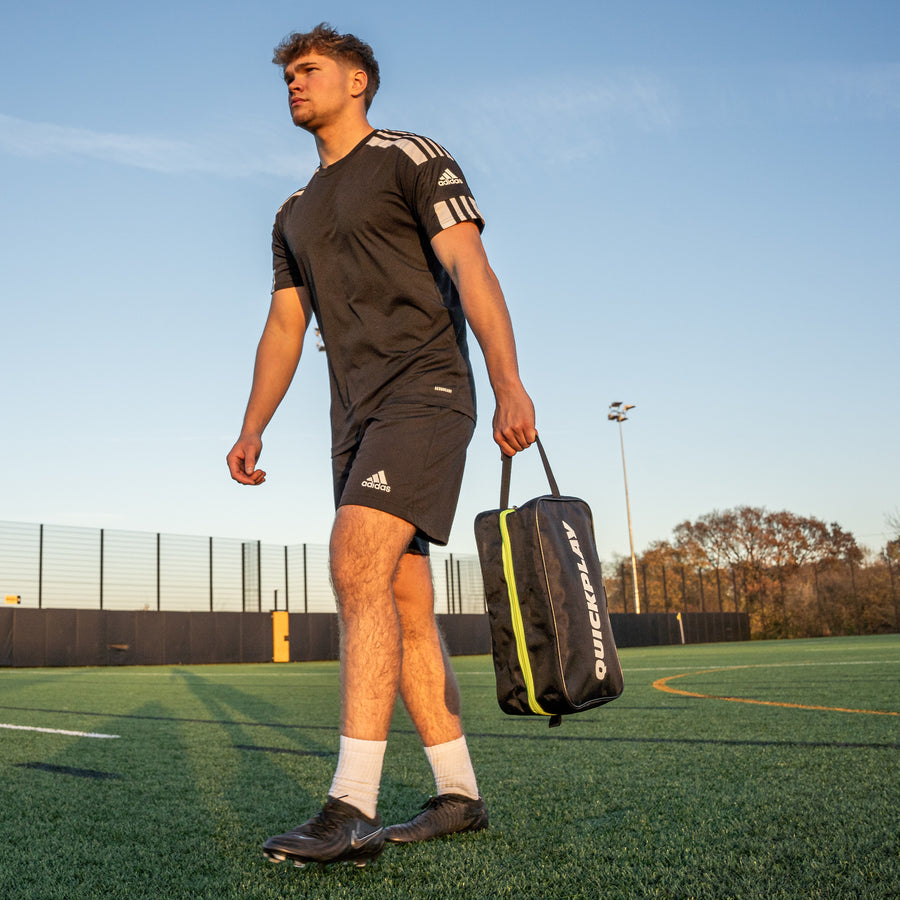 Male soccer player carrying a QUICKPLAY black cleat bag with green zipper on grass