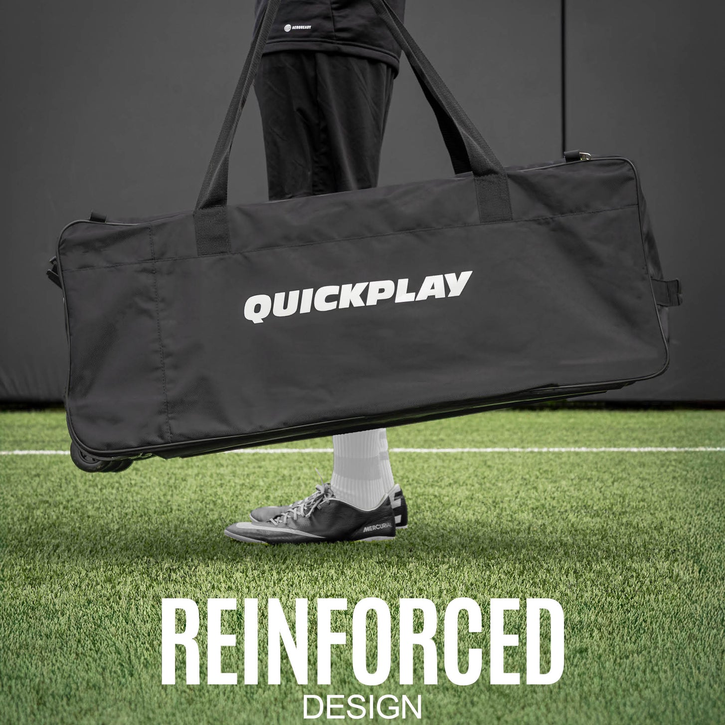 Player in cleats holding the Equipment Bag with Wheels (XL) by its handles on an artificial field, with the text "Reinforced design".