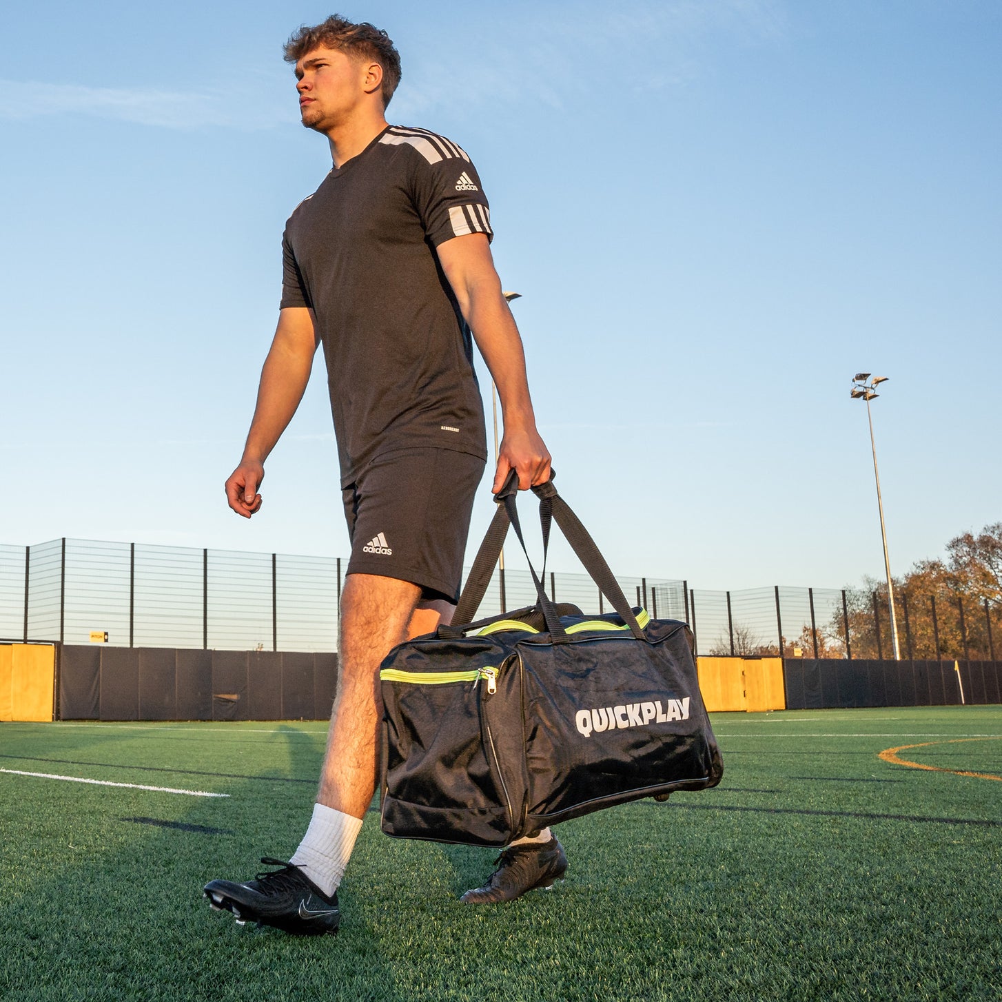 Player walking across an artificial field carrying the black Equipment Duffel Bag (M) with lime green zippers.