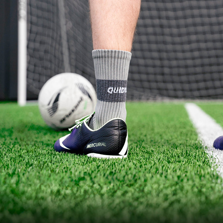 Person wearing gray socks with QUICKPLAY branding and black soccer cleats on a grass field.