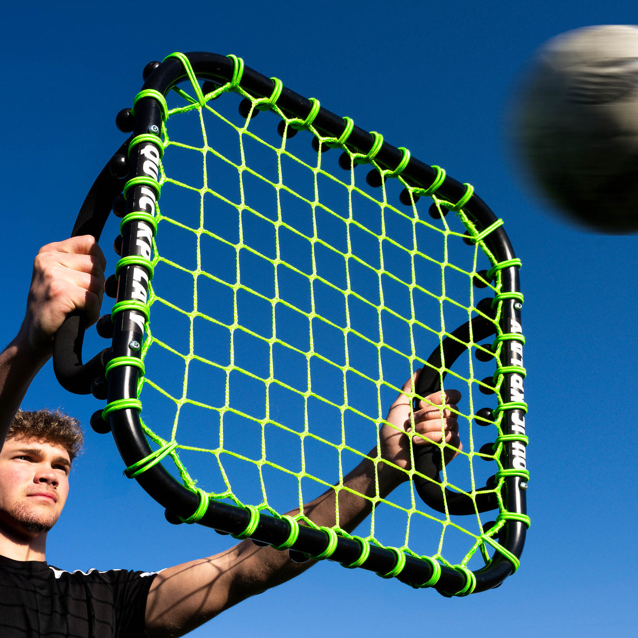 Person holding a black and green handheld rebounder against a clear blue sky