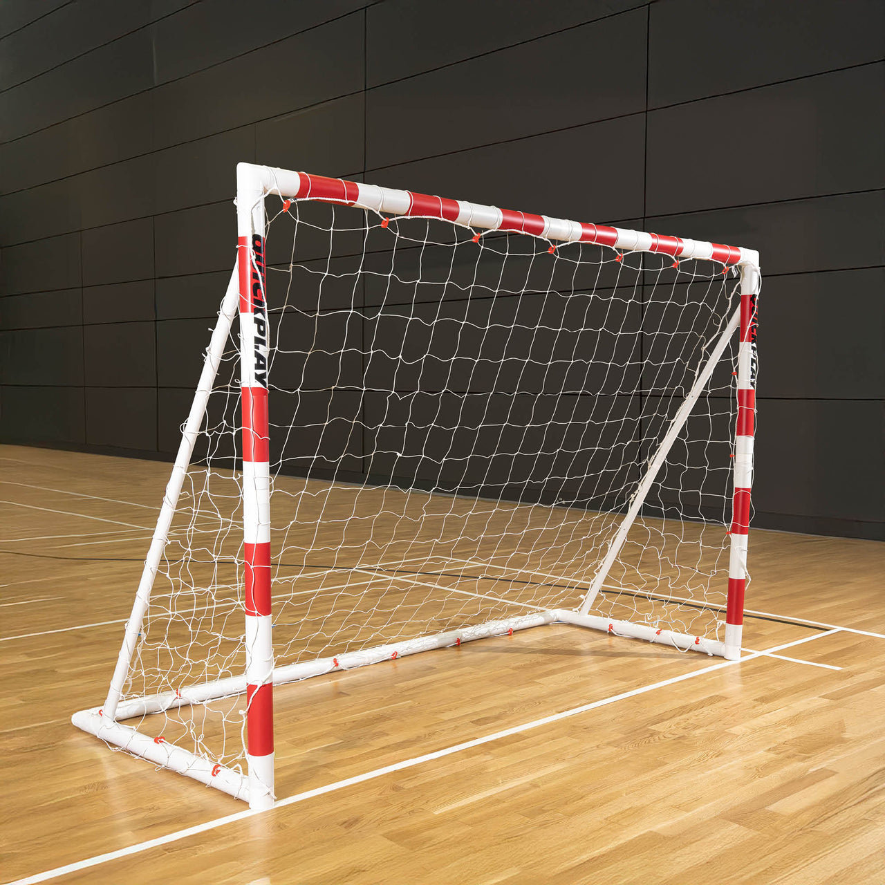 QUICKPLAY Handball goal with red and white frame on a wooden floor against a dark paneled wall.