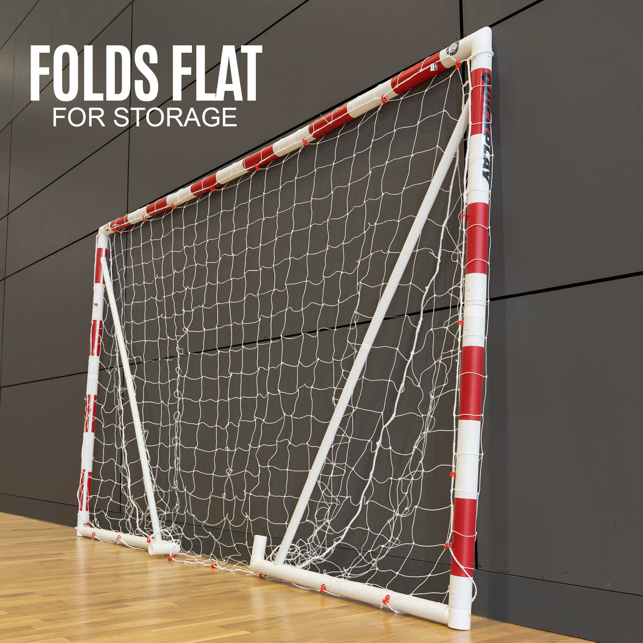 Folded QUICKPLAY Handball goal with red and white design on a wooden floor against a dark paneled wall.