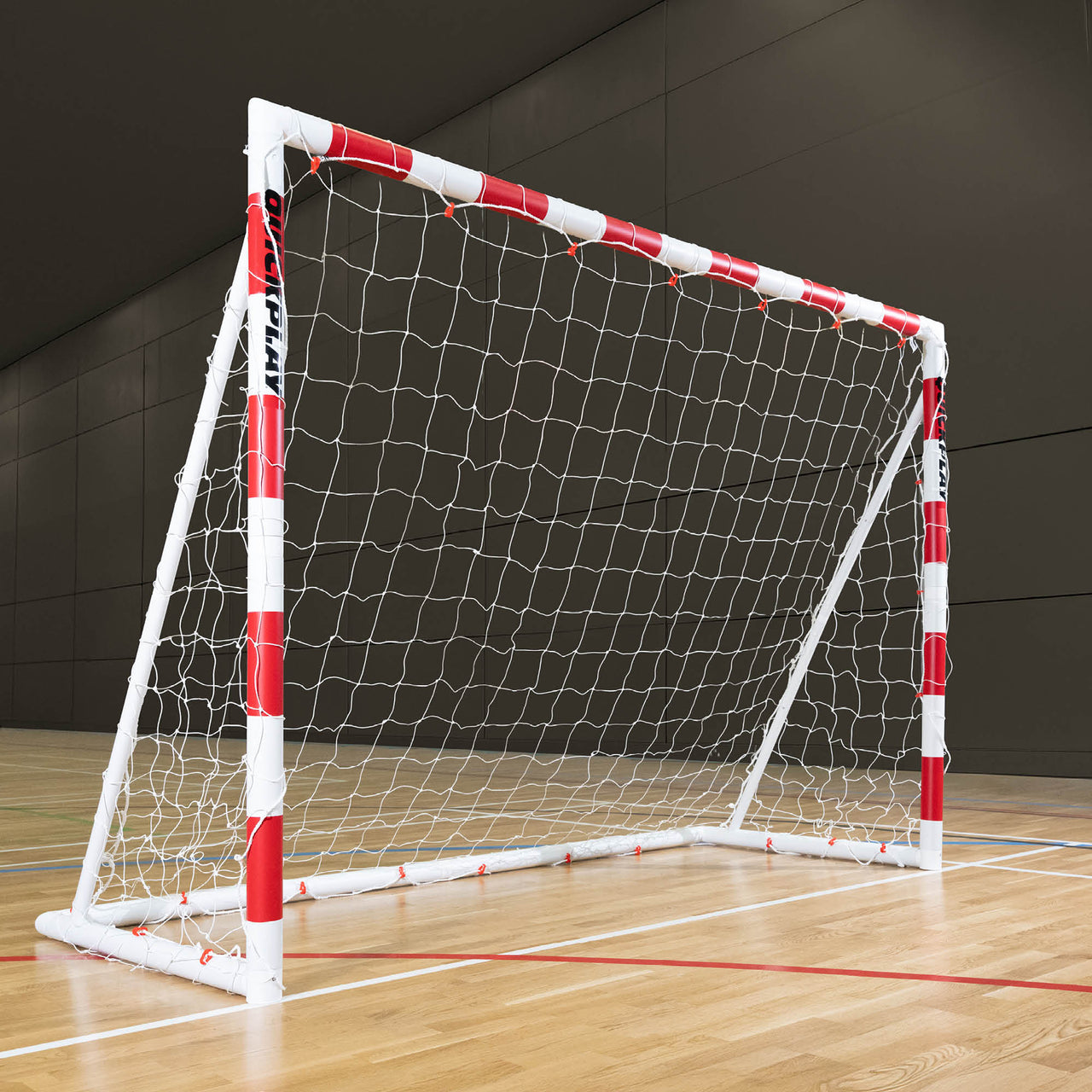 QUICKPLAY Handball goal with red and white frame on a wooden floor against a dark wall.