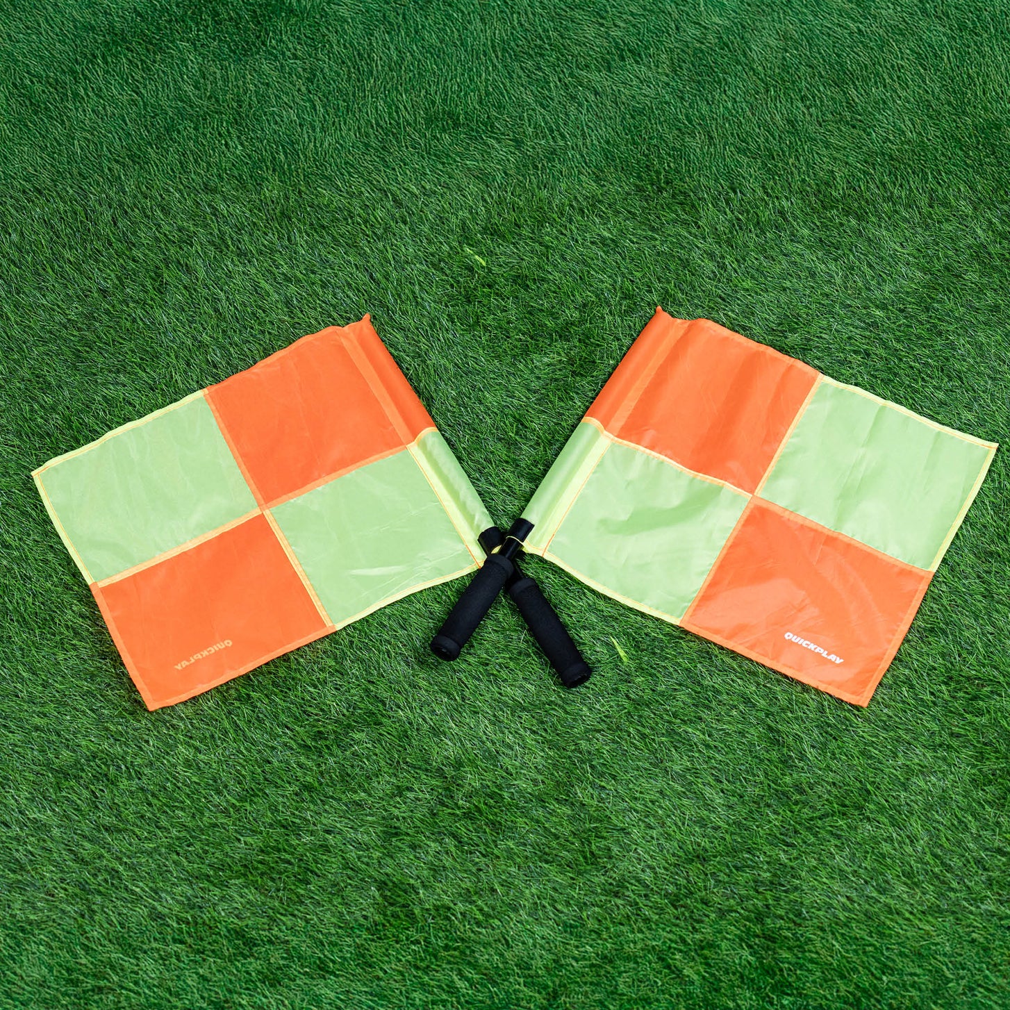 Linesman flags: two orange and yellow-green checkered flags with black handles, crossed on artificial grass.