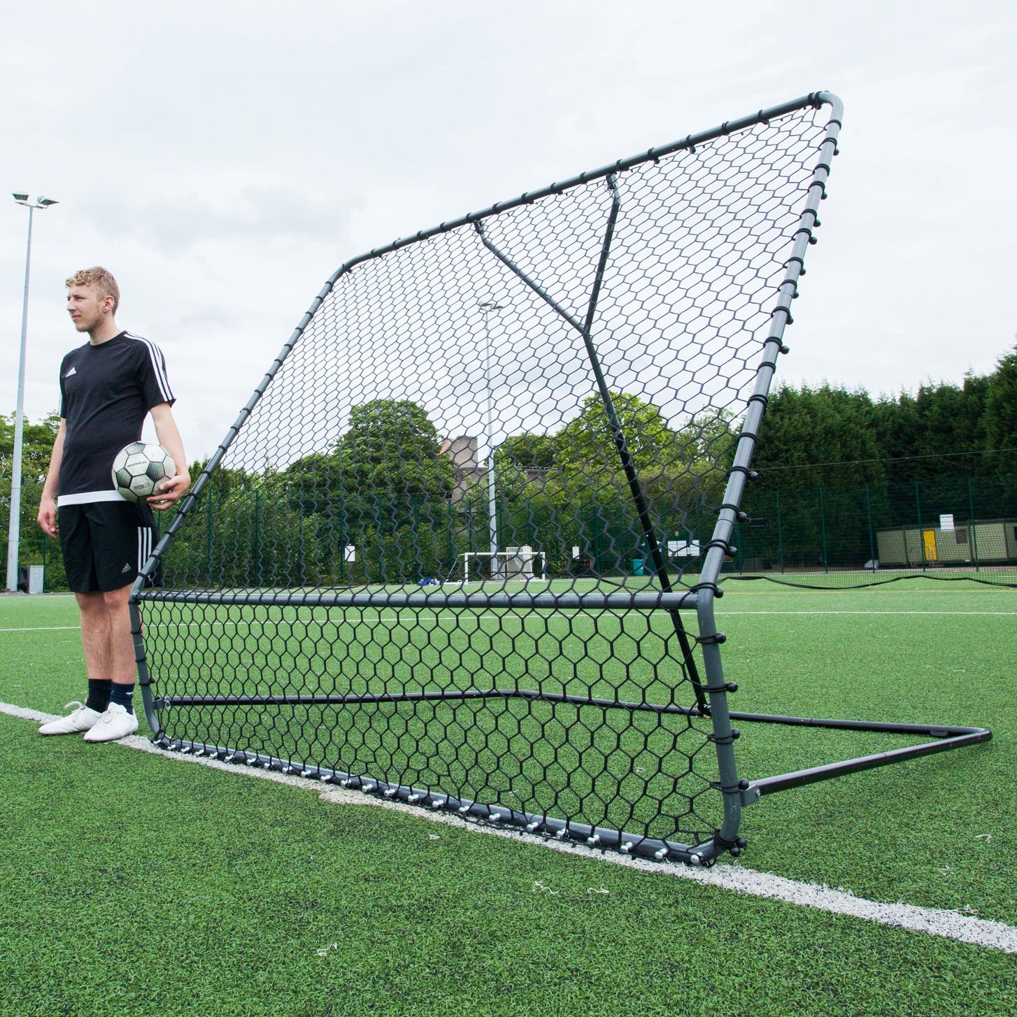 PRO Rebounder 7x7' with its black net and steel frame on an artificial field, a player with a ball standing beside it.