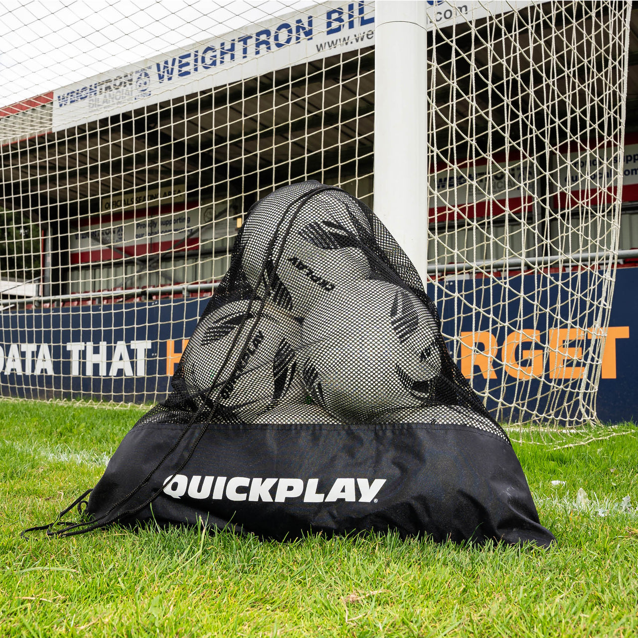 Black mesh soccer ball bag with soccer balls on a grass field in front of a goal.