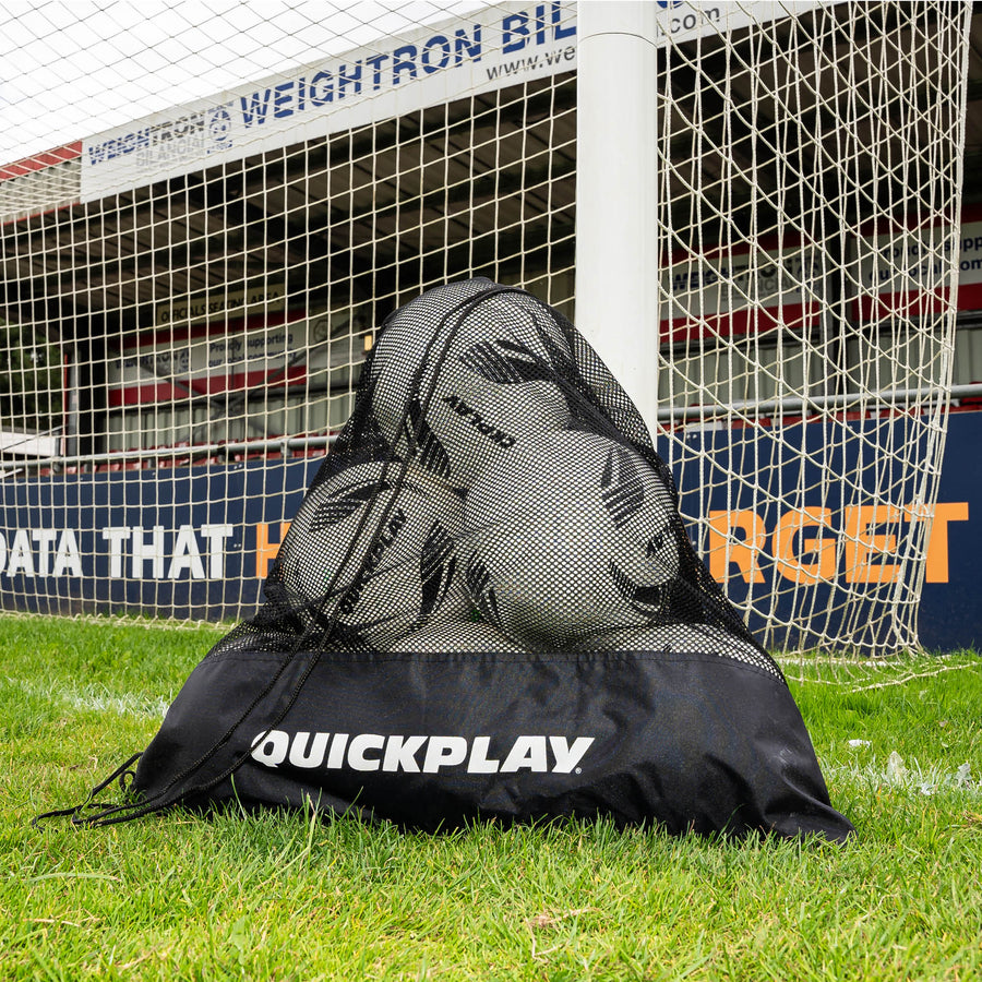 Black mesh soccer ball bag with soccer balls on a grass field in front of a goal.
