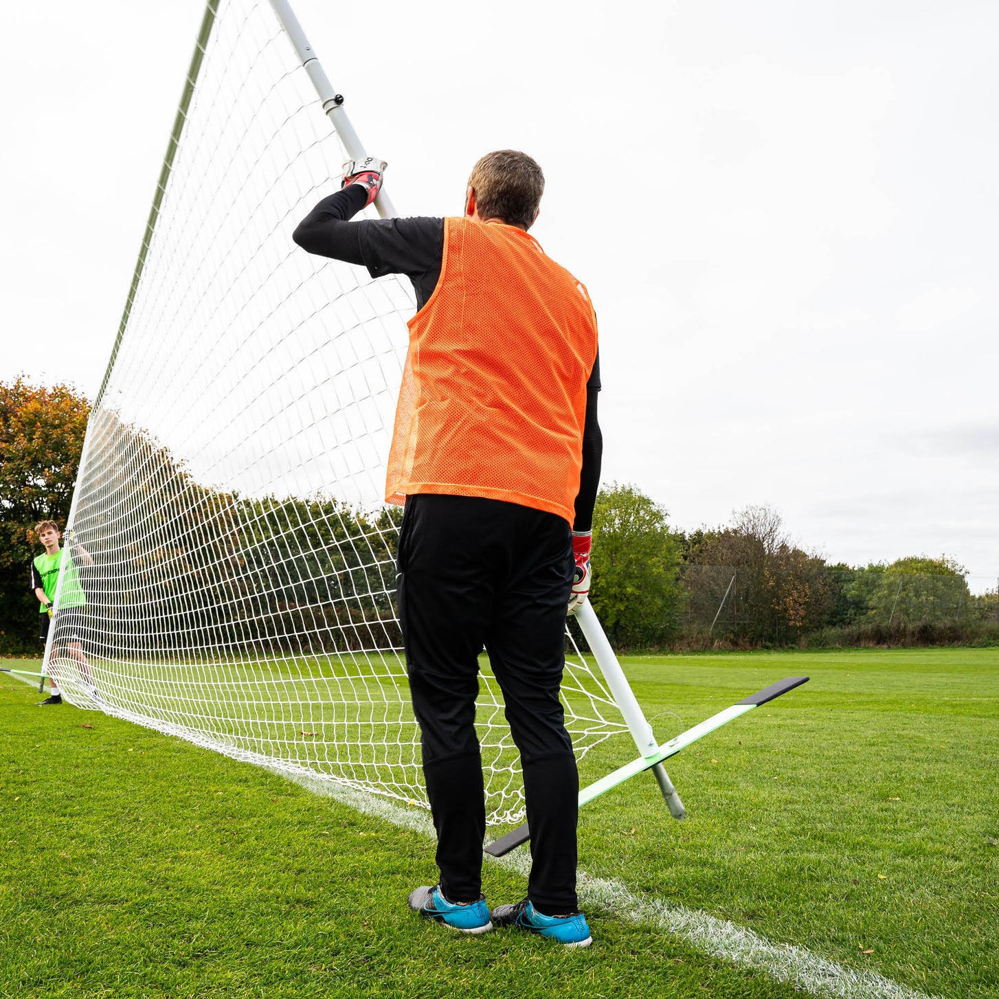 Coach in an orange bib lifting the PRO ALU Soccer Training Frame 18.5x6.5' upright on a grass field, with the base bar on the ground behind.