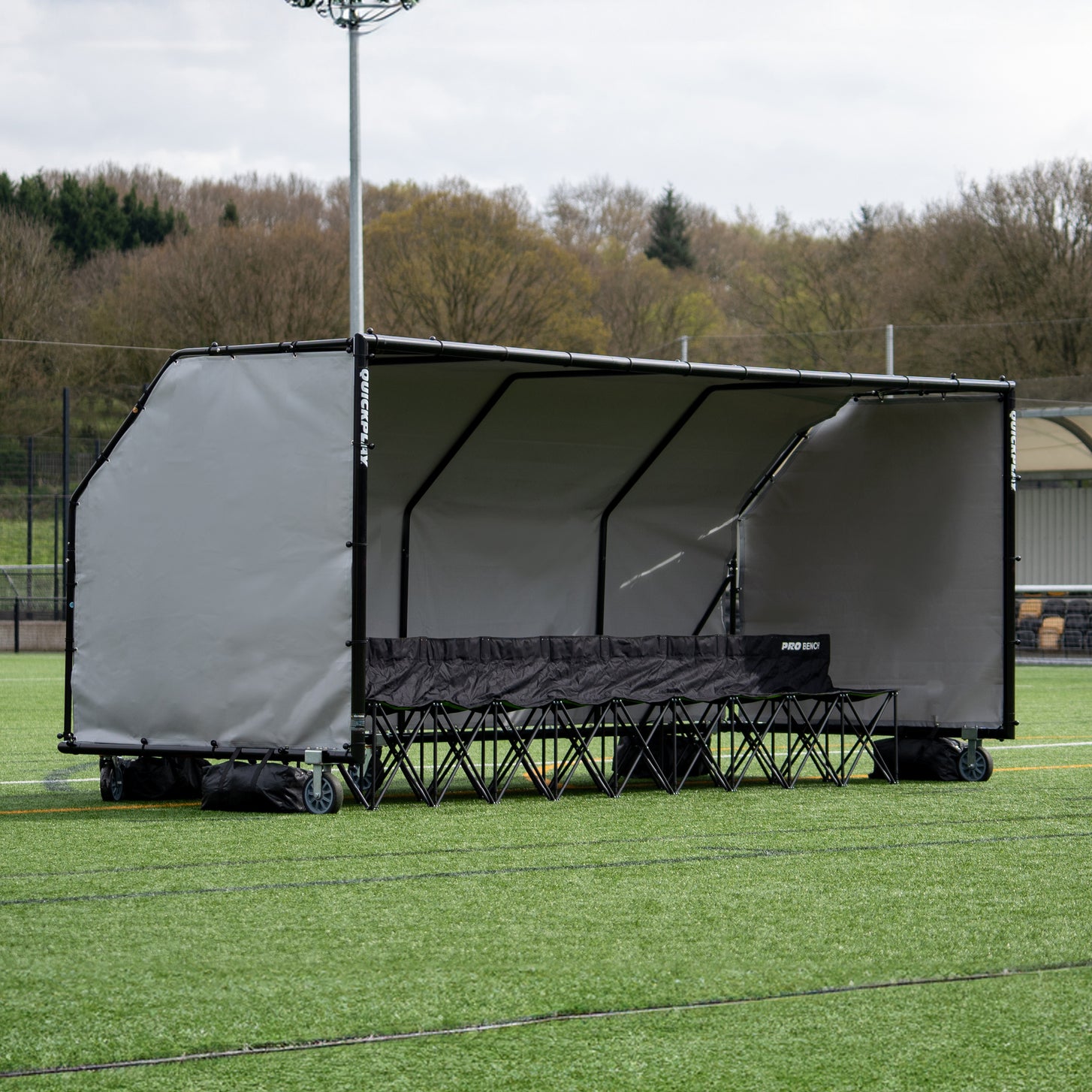 PRO ALU Club Shelter 16x6' from the other end, with a black PRO BENCH inside, on an artificial field in front of trees.