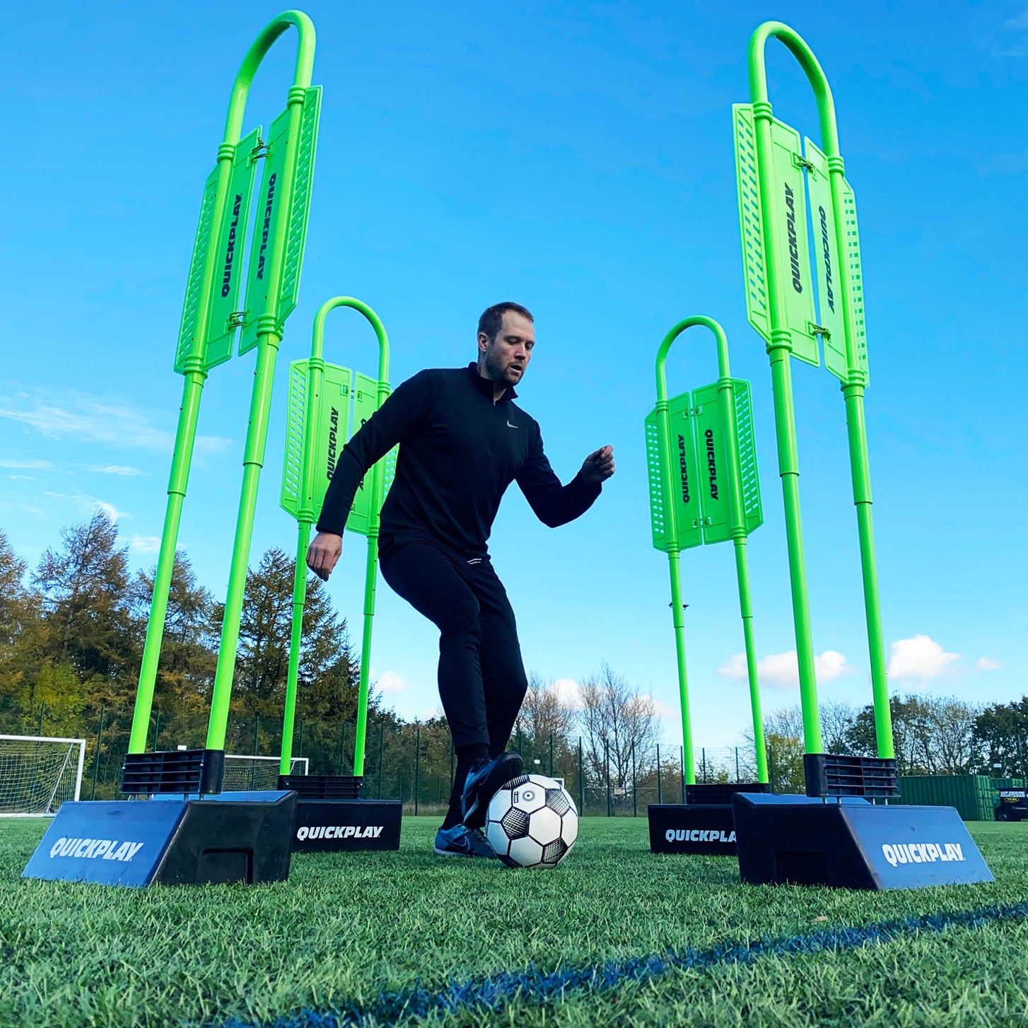Player dribbling a soccer ball between four green REPLAY Defender & Block System frames on black bases, on an artificial field.
