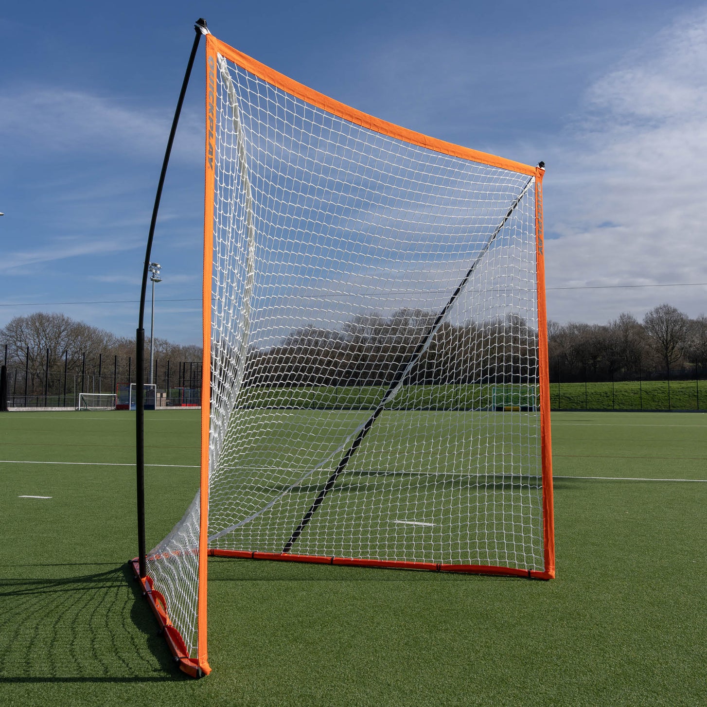 Spare lacrosse net fitted to a black frame with orange edging, standing on an artificial field.