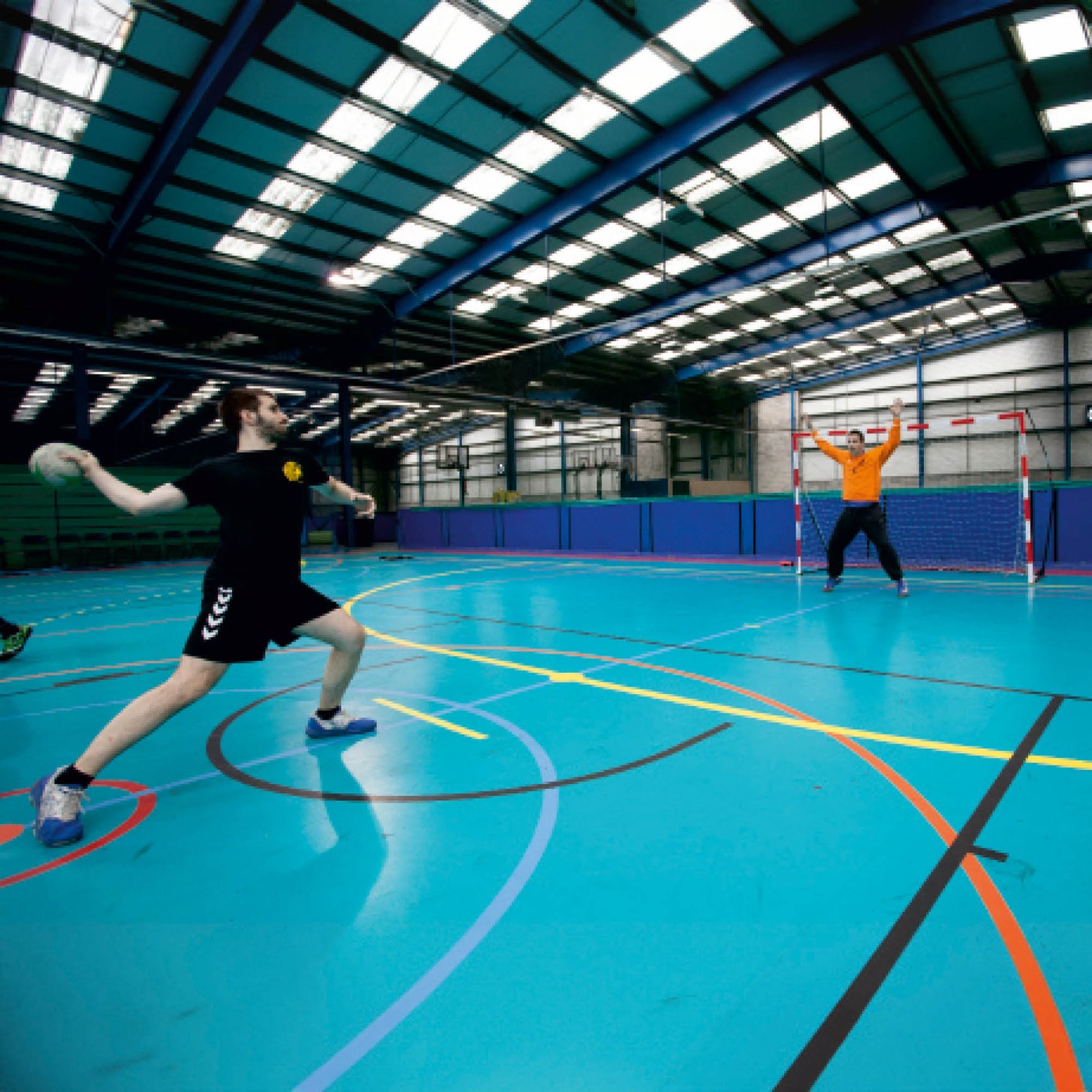 Portable Youth Handball Goal 7.9x5.6' in a sports hall as a player throws a handball toward a goalkeeper guarding the goal.