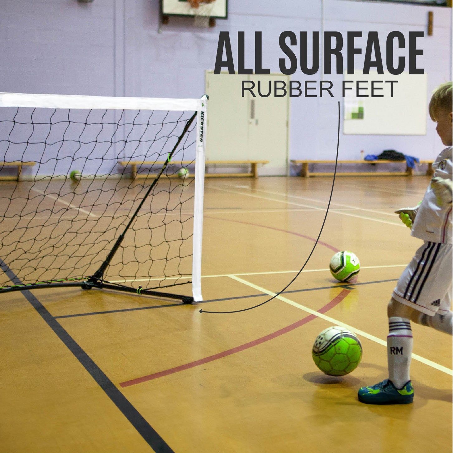Child shooting at a KICKSTER Elite goal on a sports hall floor, with the text "All surface rubber feet" pointing to its base.