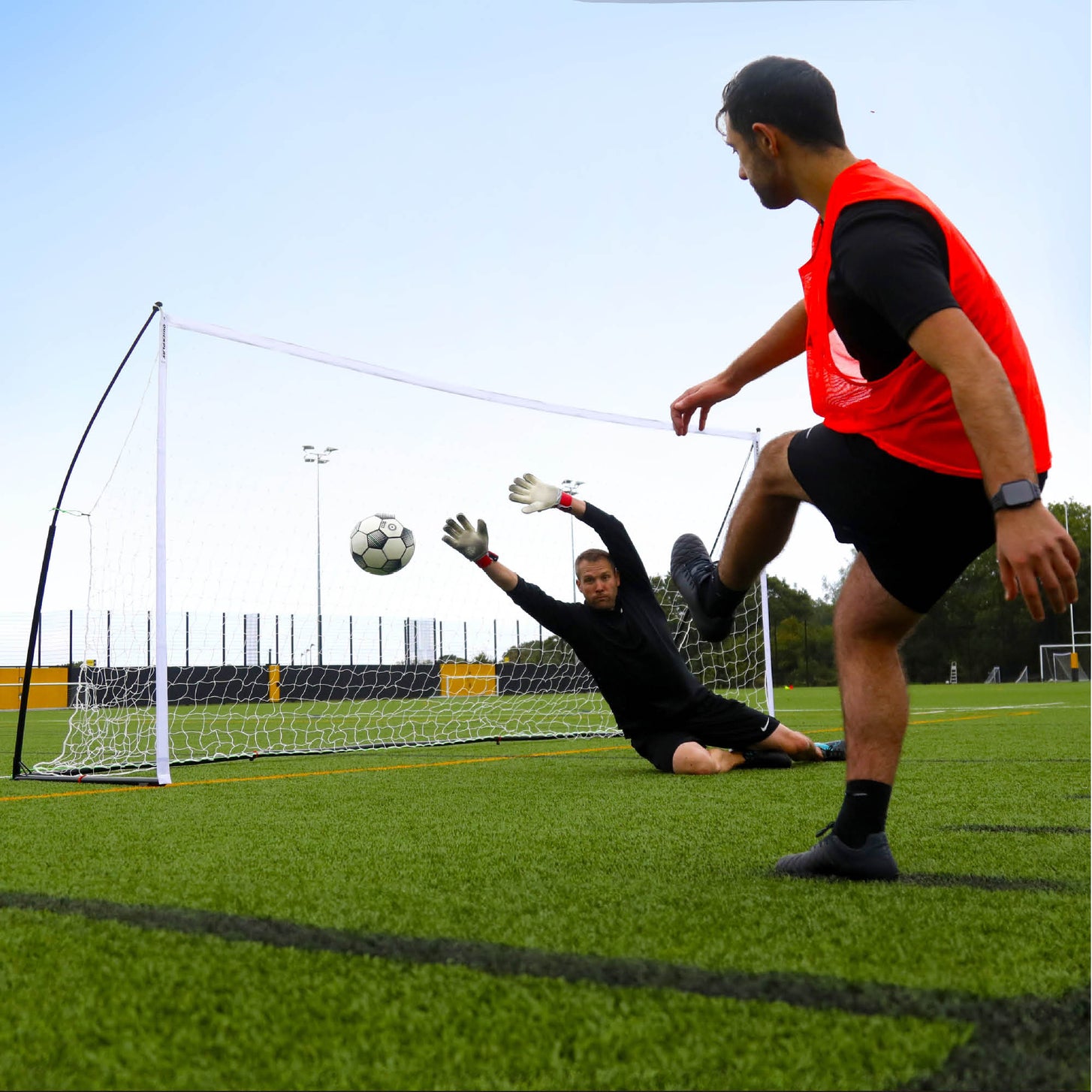 Goalkeeper diving as a player in an orange bib shoots at the KICKSTER Elite 18.5x6.5' goal on an artificial field.