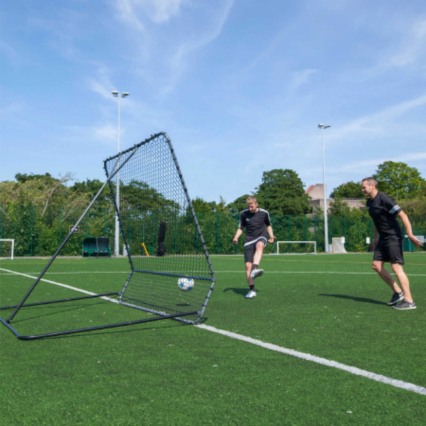 PRO Rebounder 7x7': two players passing a ball off it in turn on an artificial field under blue sky.