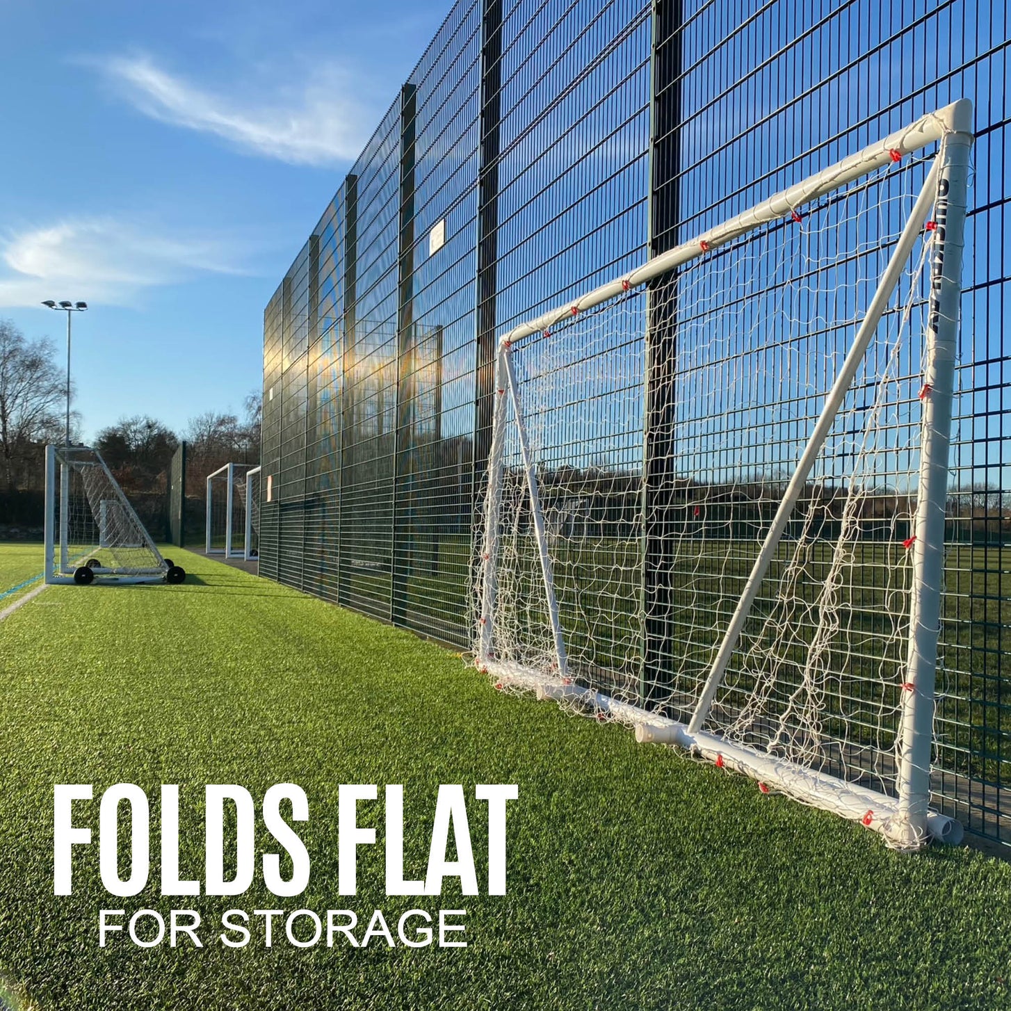 Q-FOLD Folding Soccer Goal 16x7' folded flat against a tall mesh fence beside an artificial field, with the text "Folds flat for storage".