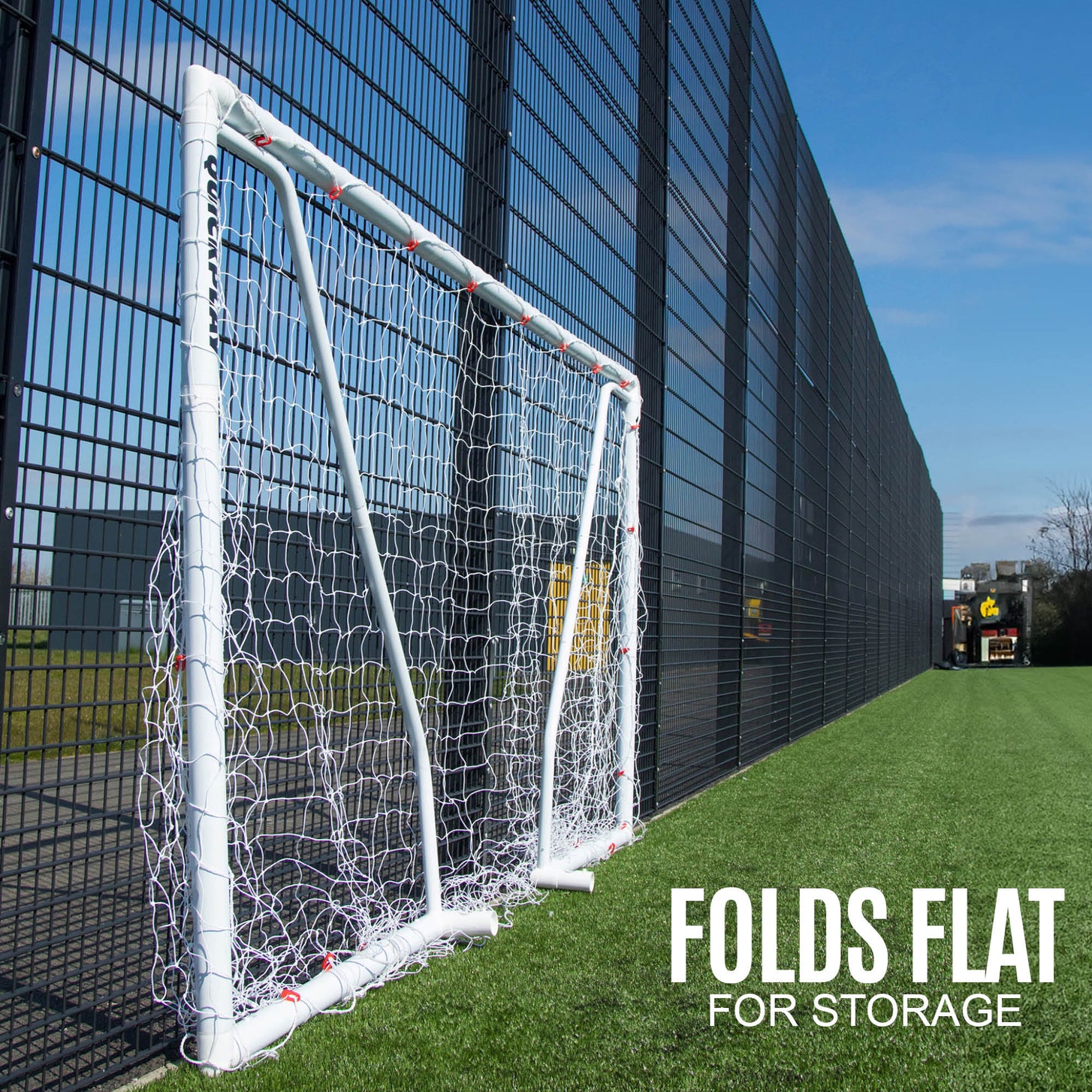 Q-FOLD Match futsal goal 9.8x6.5' folded flat against a tall fence on an artificial field, with the text "Folds flat for storage".