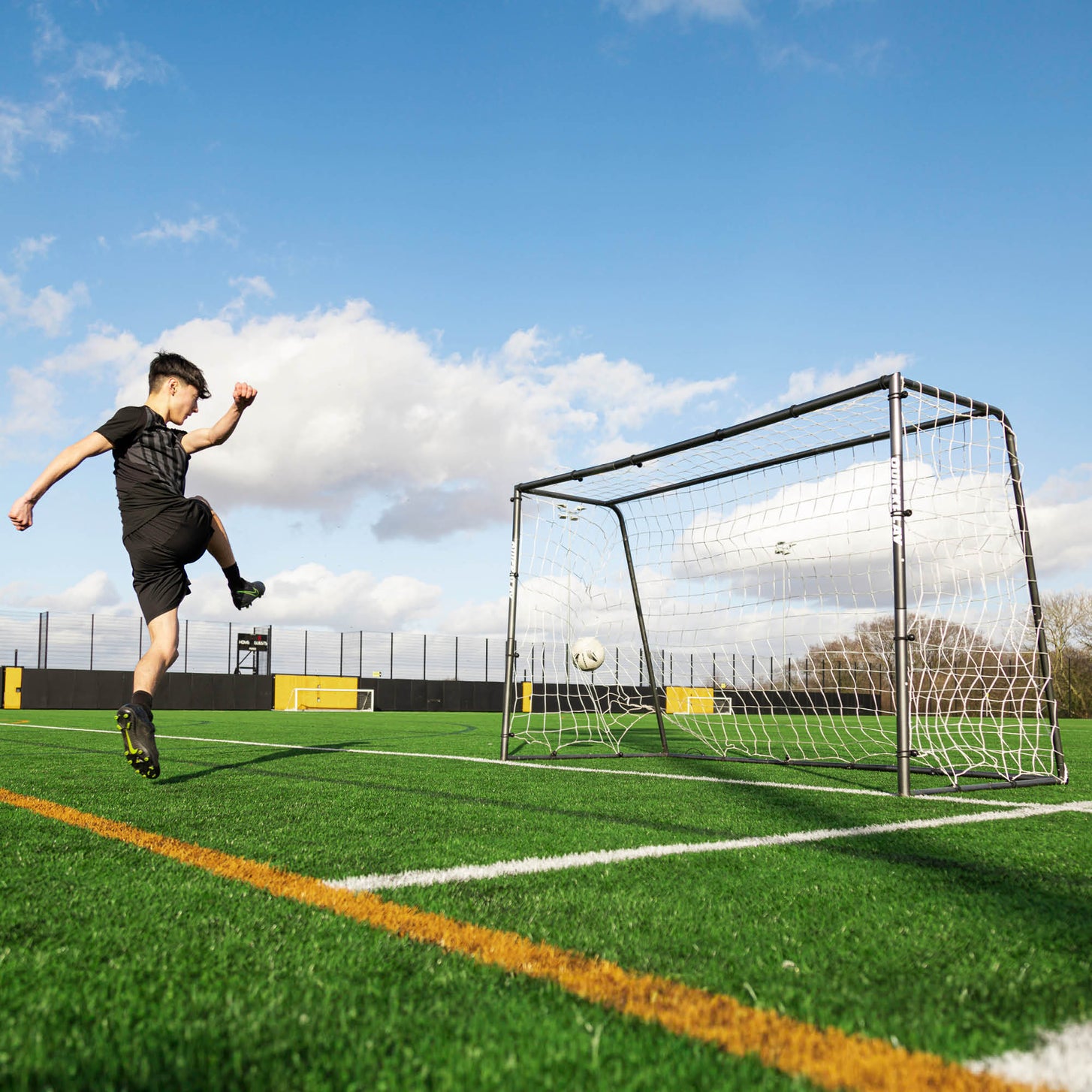 Player in black volleying a soccer ball into the GS Series Soccer Goal 6x4' on a sunny artificial field.