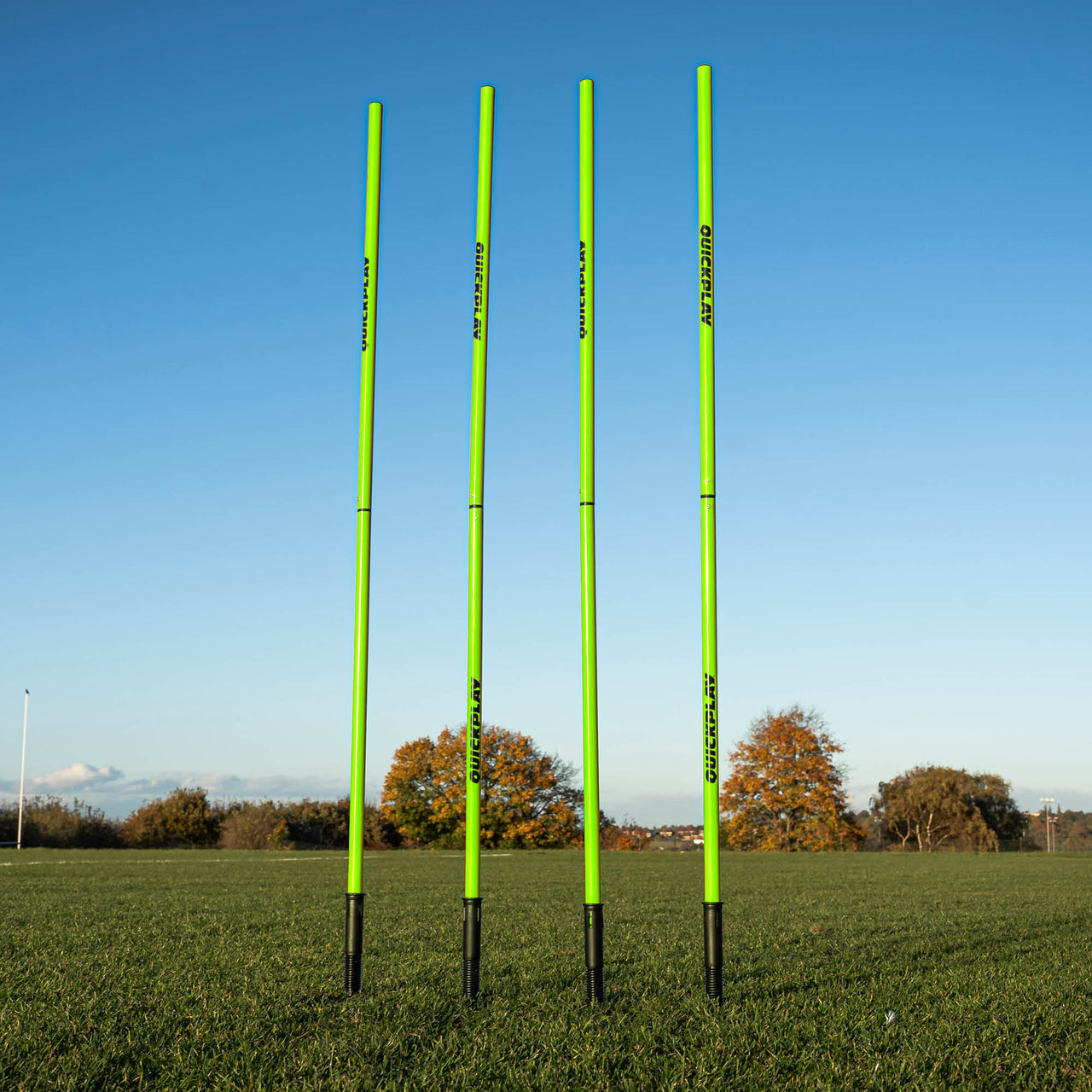 A set of four green training poles with sprung stake bases on a grass field, designed for speed and agility training.