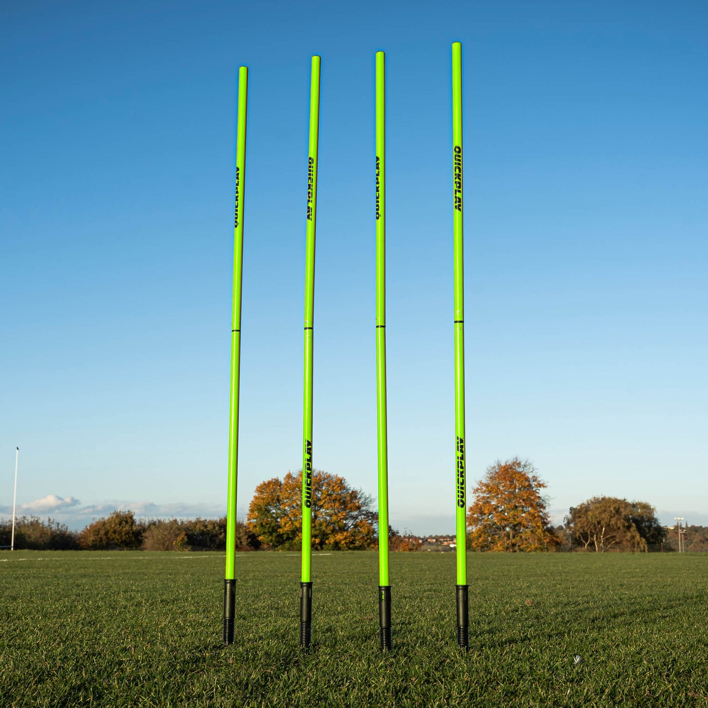 A set of four green training poles with sprung stake bases on a grass field, designed for speed and agility training.