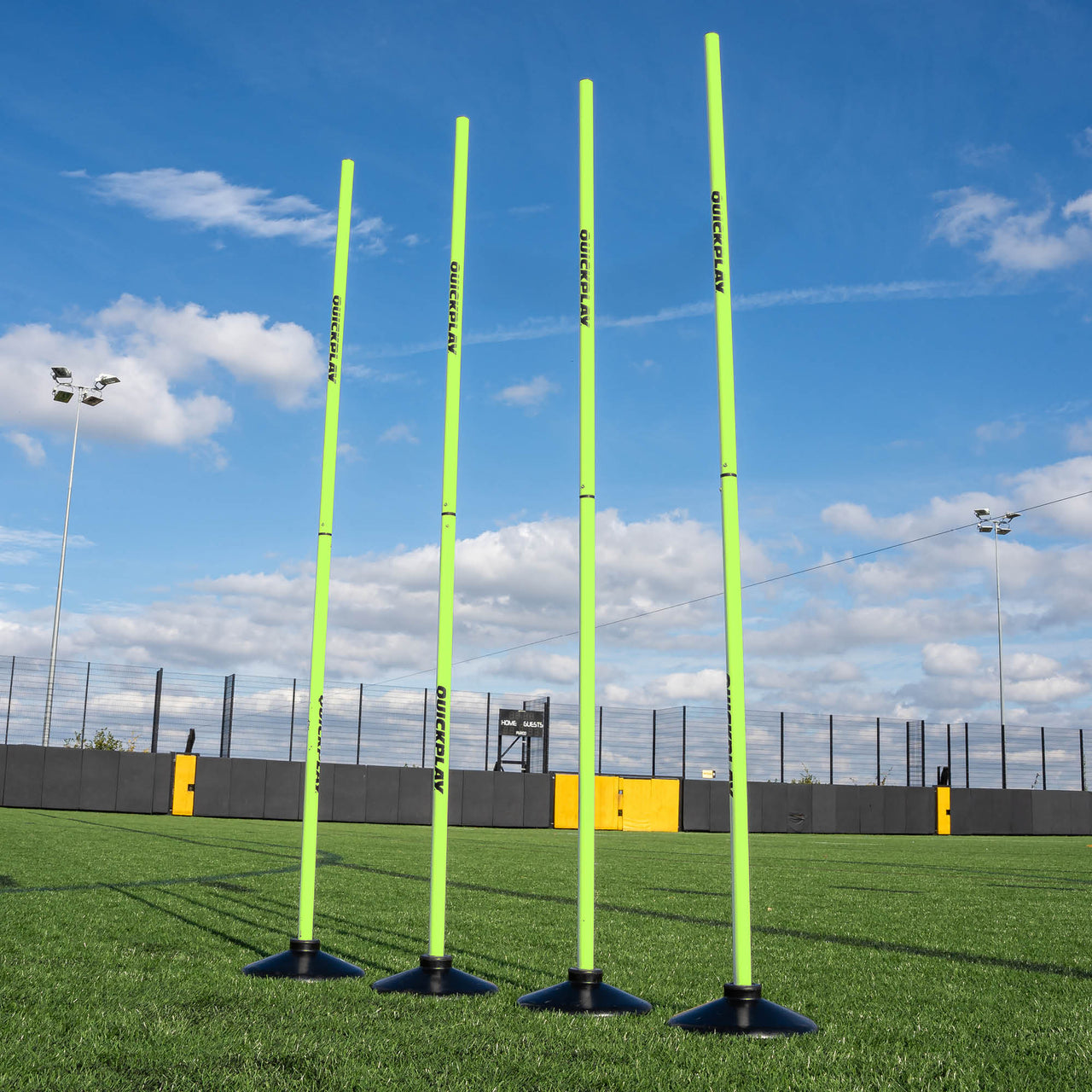 Four bright green training poles with weighted bases on a sports field with a blue sky and clouds in the background.