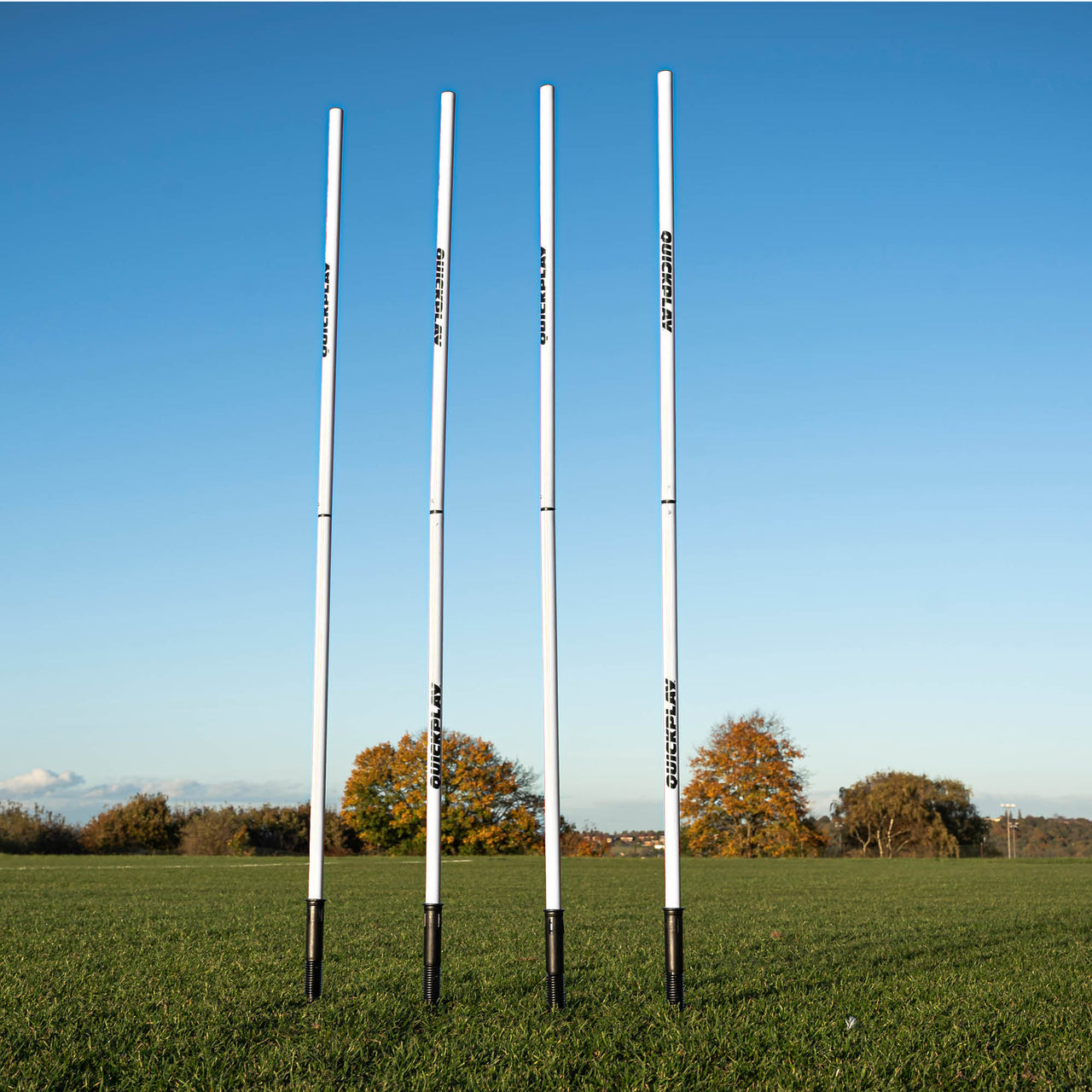 A set of four white training poles with sprung stake bases on a grass field, designed for speed and agility training.