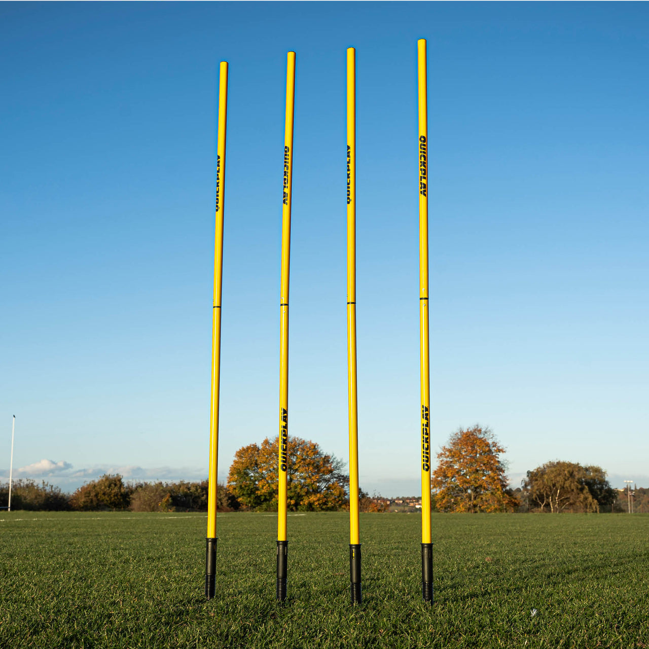 A set of four yellow training poles with sprung stake bases on a grass field, designed for speed and agility training.