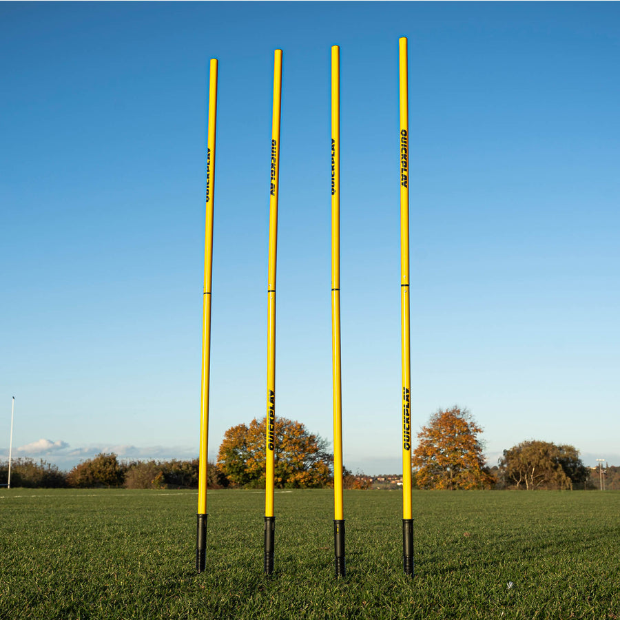 A set of four yellow training poles with sprung stake bases on a grass field, designed for speed and agility training.