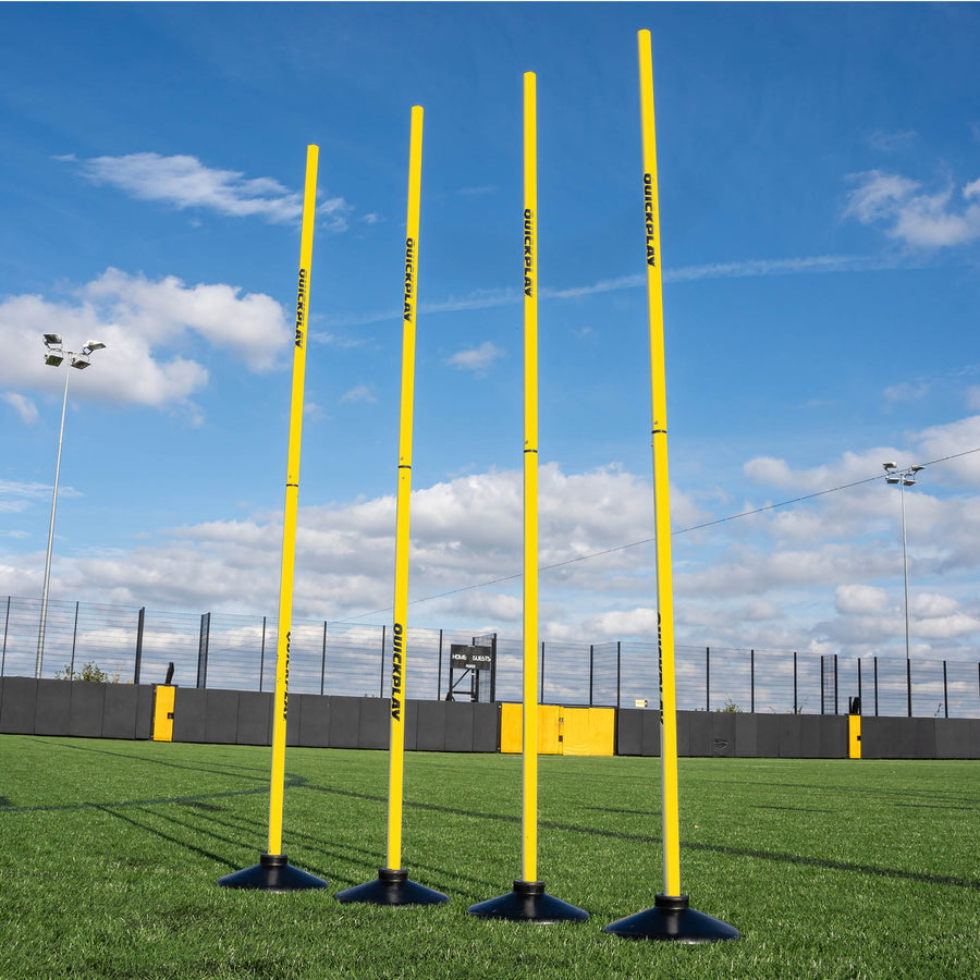 A set of four yellow training poles with weighted bases on a grass field, designed for speed and agility training.