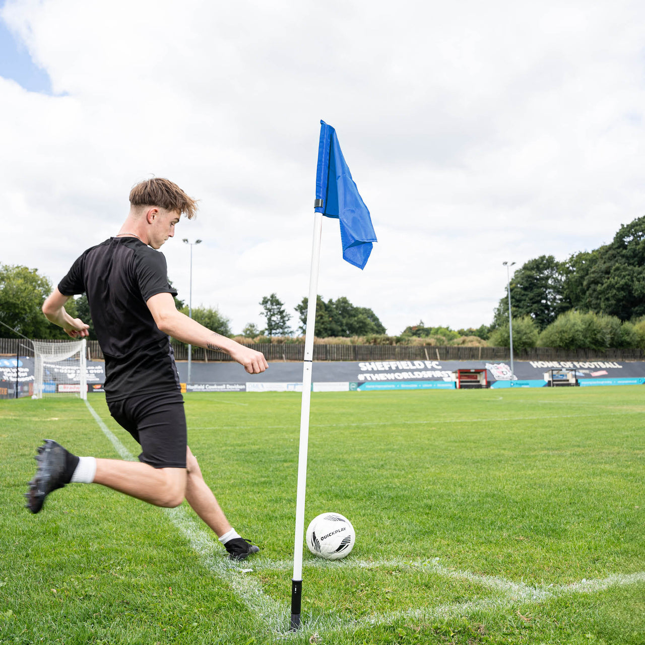 A soccer corner flag in blue attached to a pole with a sprung ground stake, set against an outdoor grass pitch background.