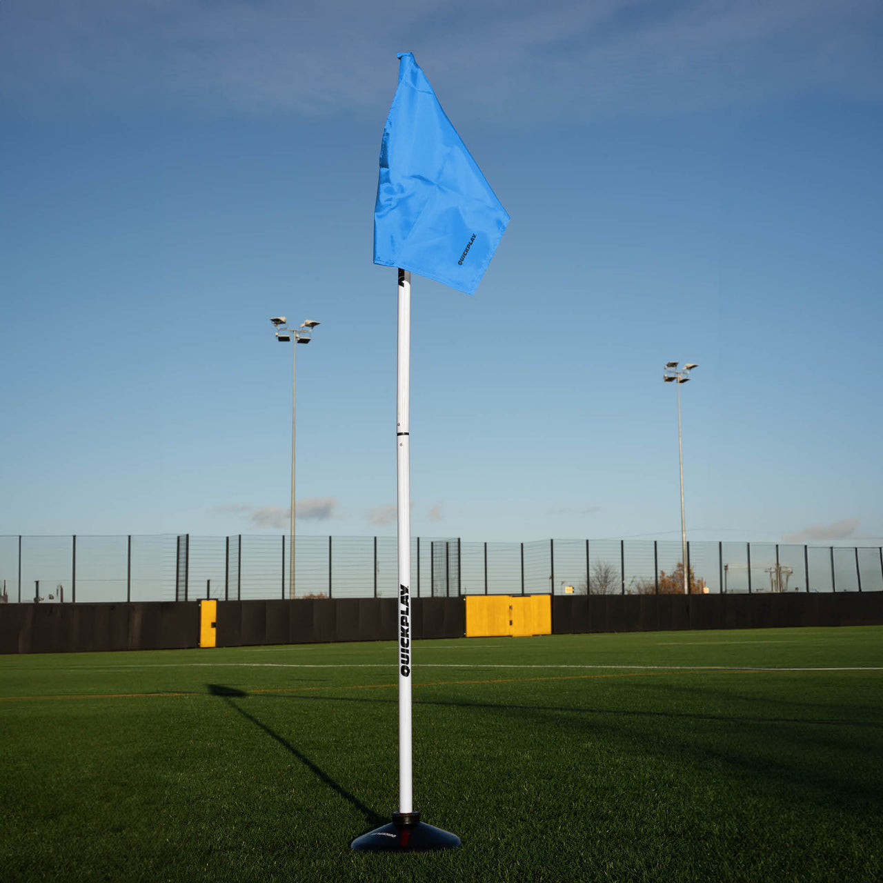 A soccer corner flag in blue attached to a pole with a weighted base, set against an outdoor grass pitch background.