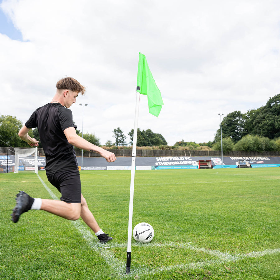 A soccer corner flag in green attached to a pole with a sprung ground stake, set against an outdoor grass pitch background.
