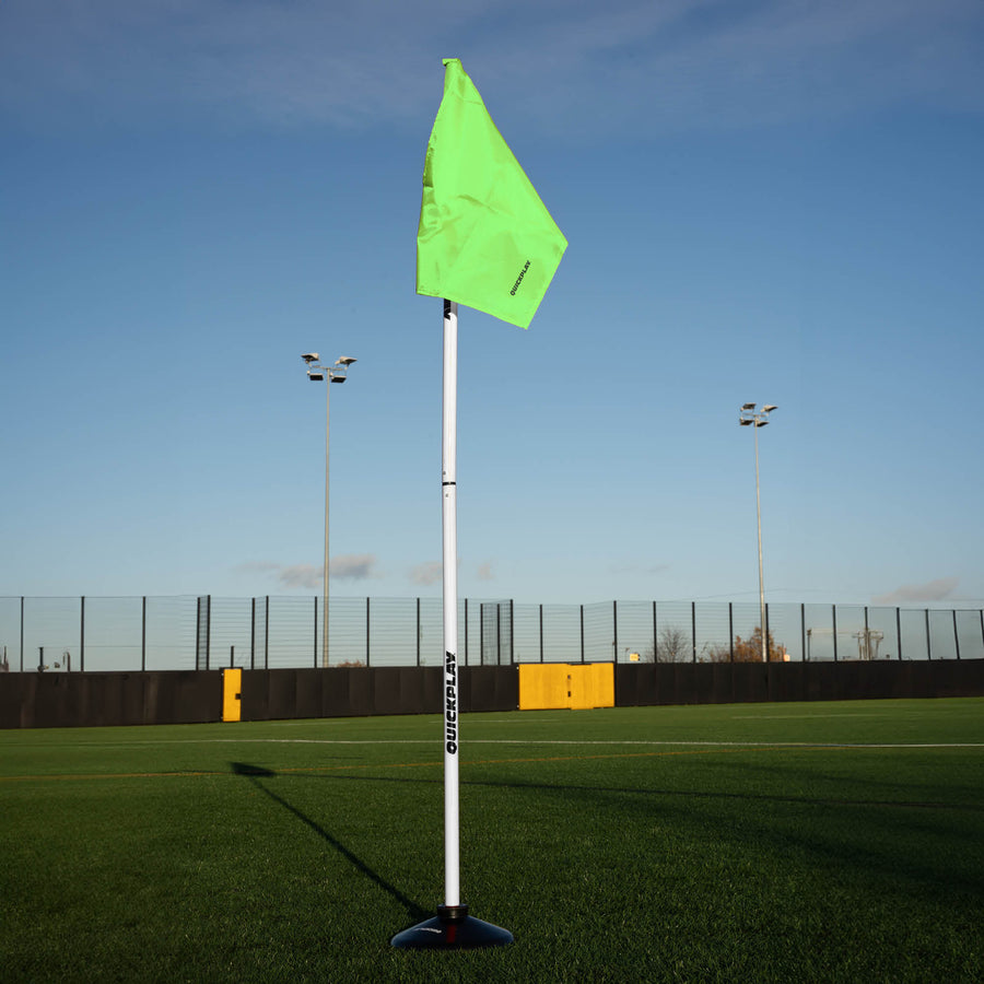 A soccer corner flag in green attached to a pole with a weighted base, set against an outdoor grass pitch background.