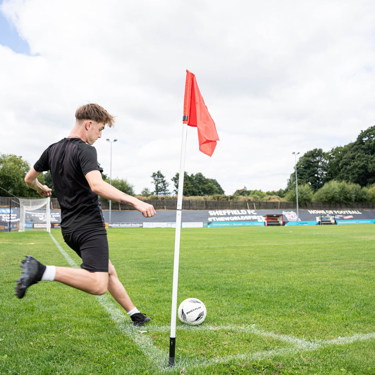 A soccer corner flag in red attached to a pole, set against an outdoor grass pitch background.