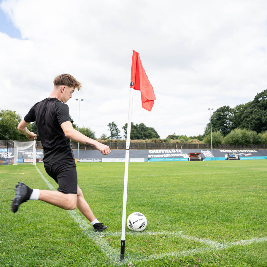 A soccer corner flag in red attached to a pole, set against an outdoor grass pitch background.