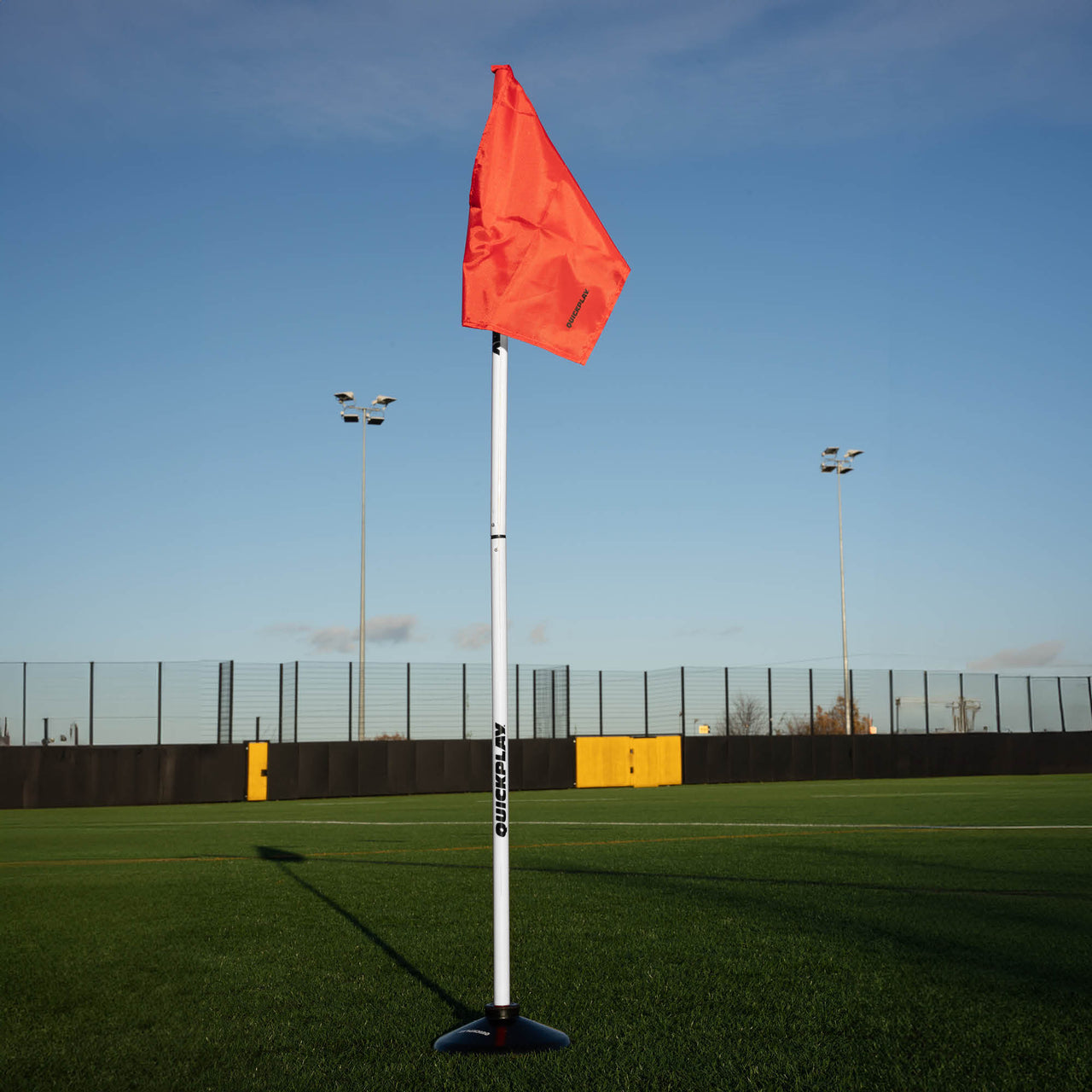 A soccer corner flag in red attached to a pole with a weighted base, set against an outdoor grass pitch background