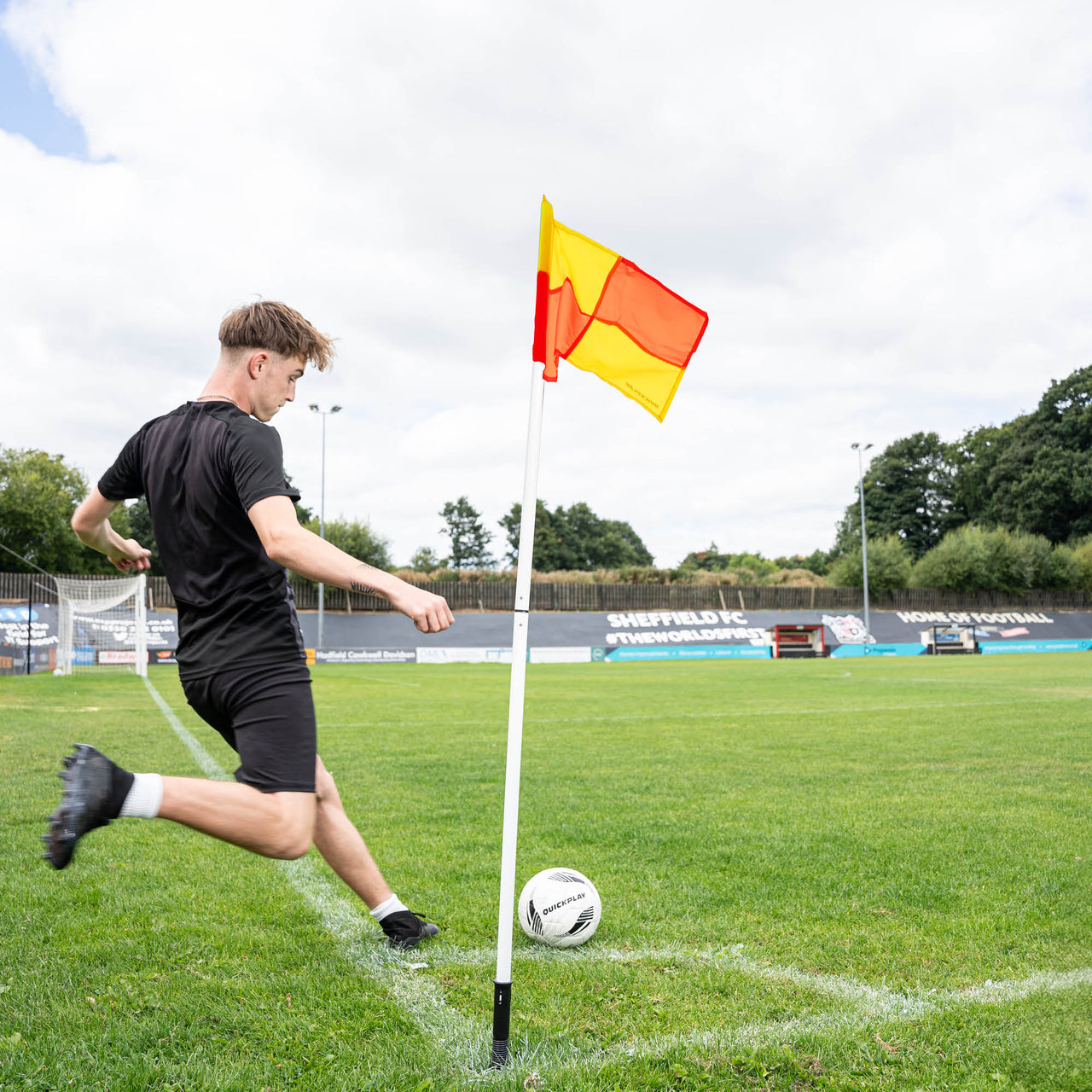 A soccer corner flag in yellow & orange attached to a pole, set against an outdoor grass pitch background.