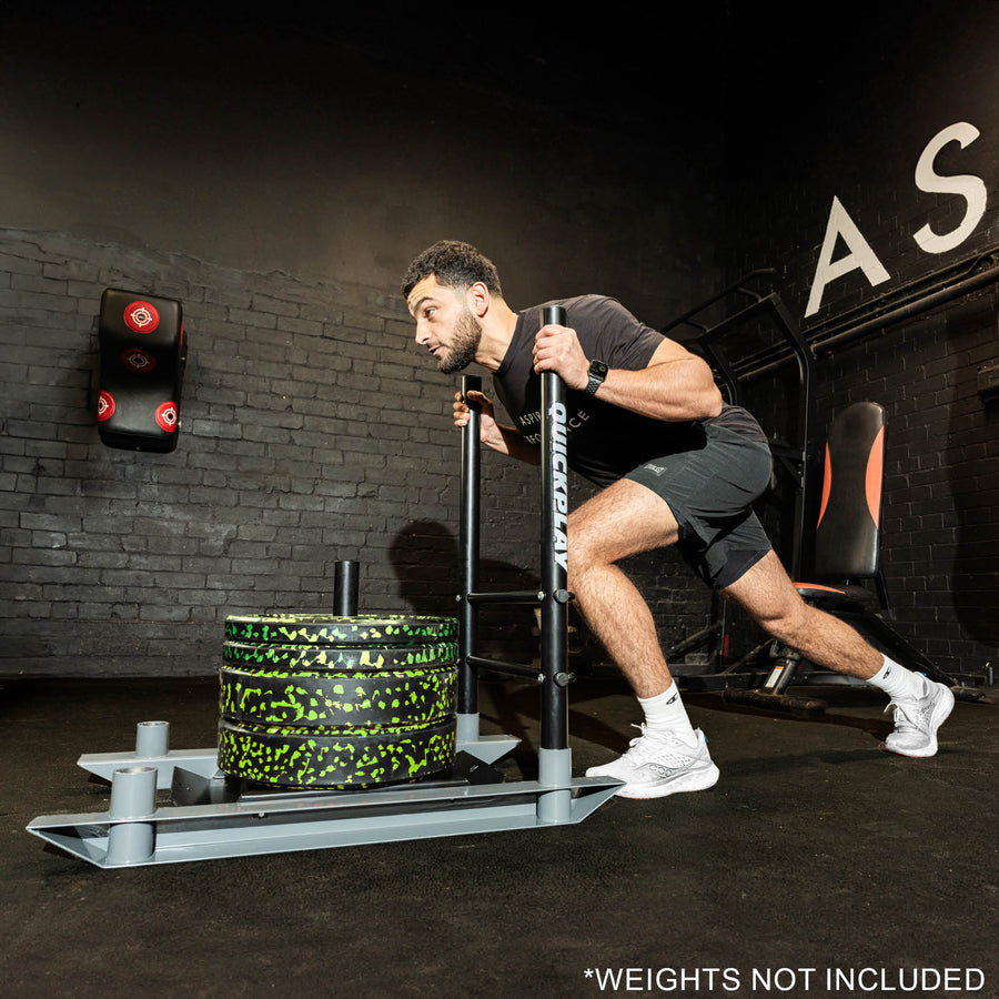 A person exercising with a weighted training sled in a gym setting. The sled has a black and green color scheme, and the person is using it for a lower body strength exercise.