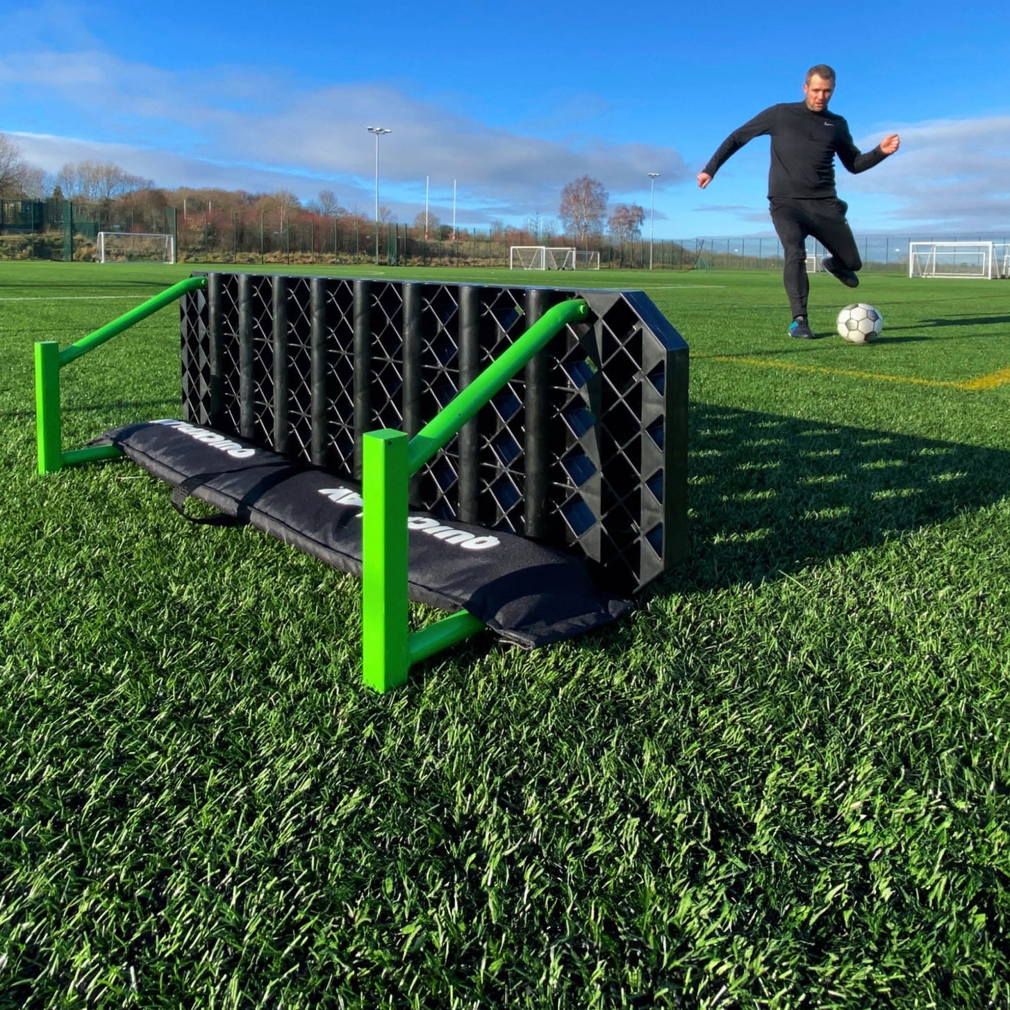 Player running in to strike a soccer ball toward the REPLAY Station XL, seen from behind the board and its sandbag.