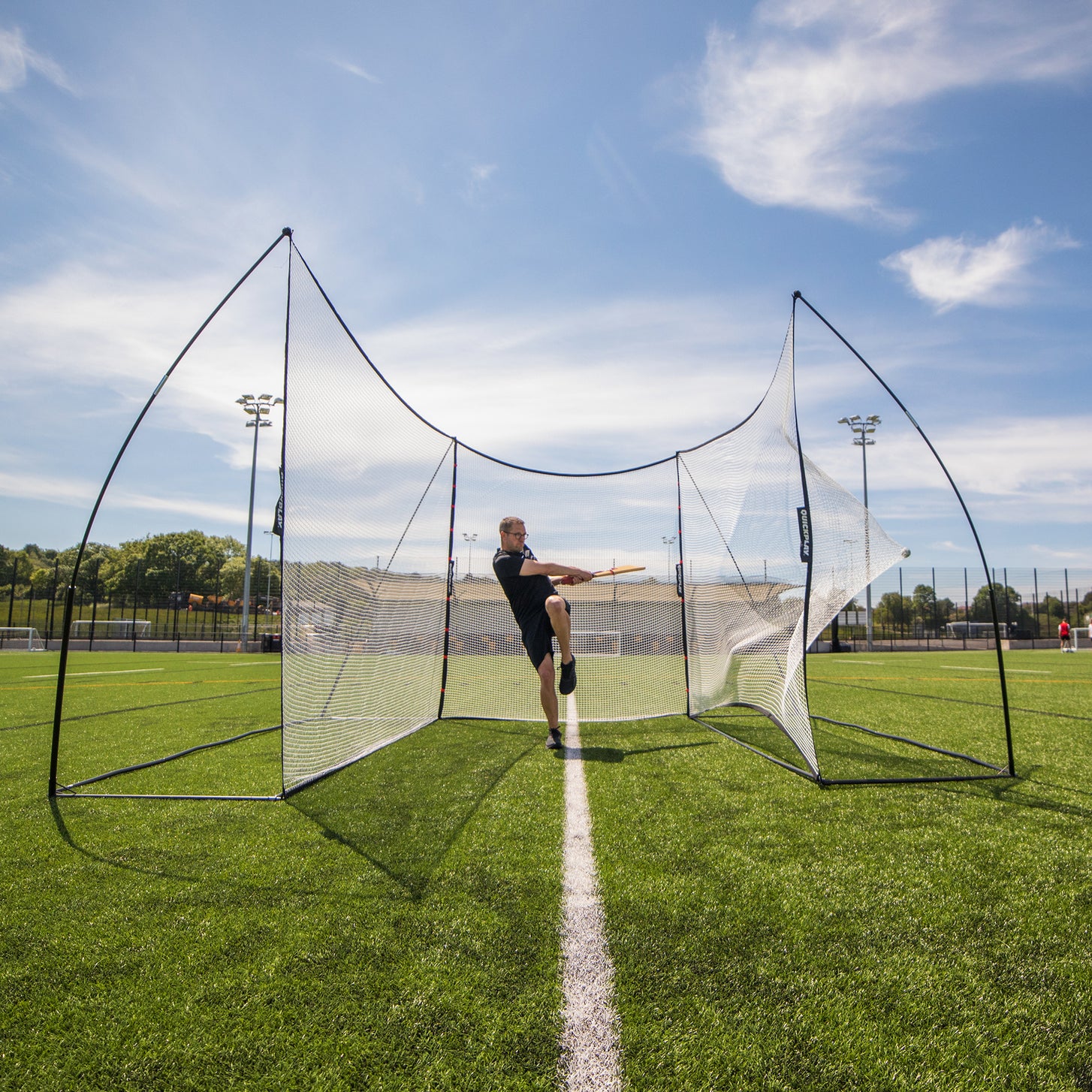 Man mid-swing with an orange bat inside the MEGA NET XL Multi Sport Backstop Net 32x9' on an artificial field.