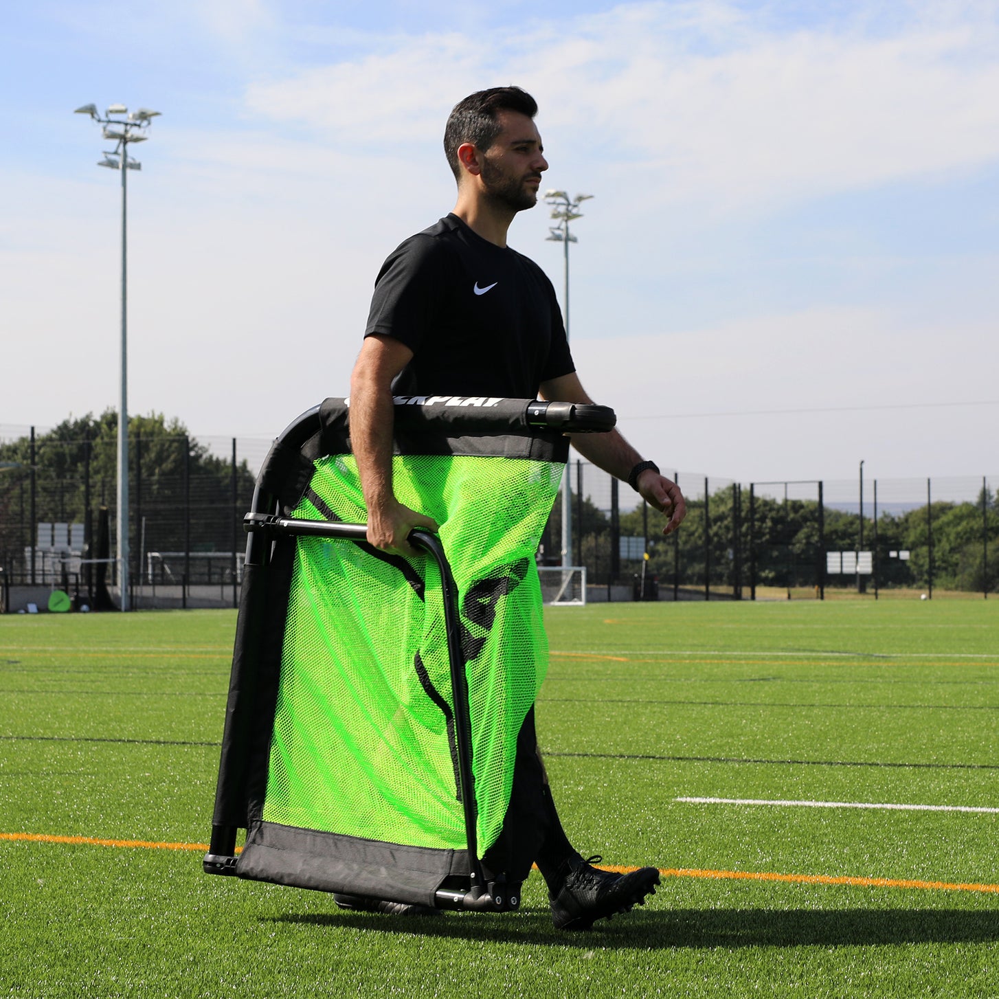 Man carrying the folded PRO ALU Training Soccer Goal 5x3' under one arm across an artificial field.