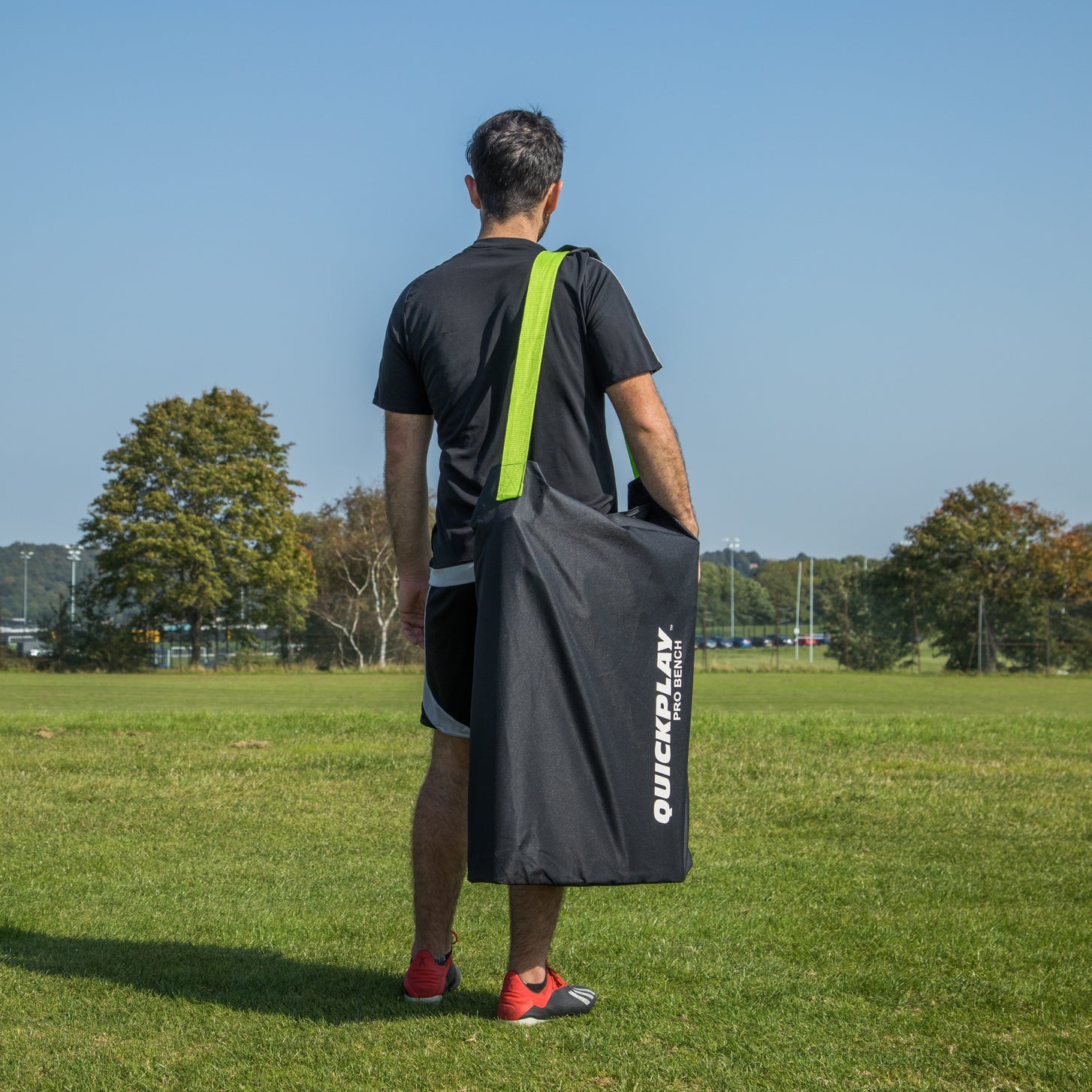 Man seen from behind carrying the PRO Bench Shelter in a black QUICKPLAY PRO BENCH bag with a green strap on a grass field.