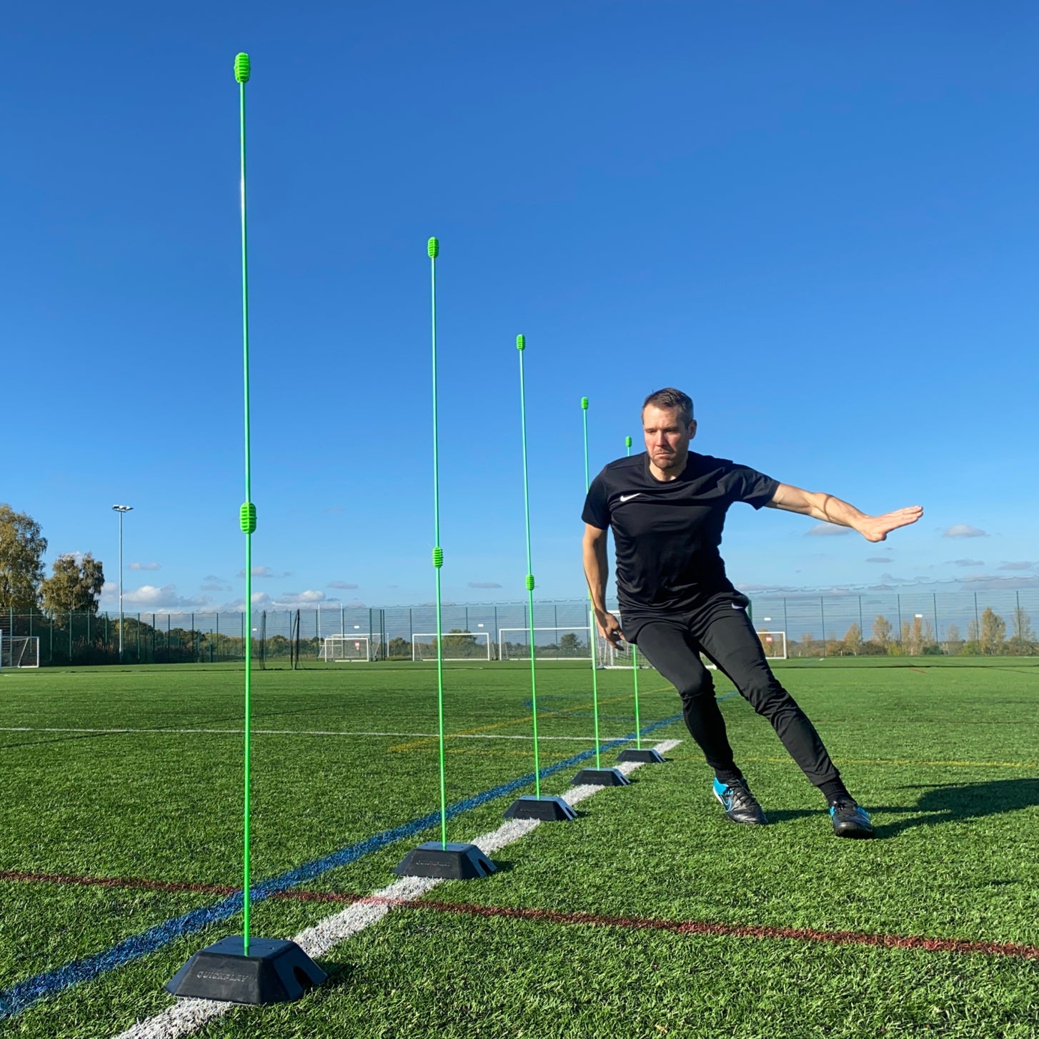 Base Weights holding green agility poles upright in a line as a man moves sideways past them on an artificial field.