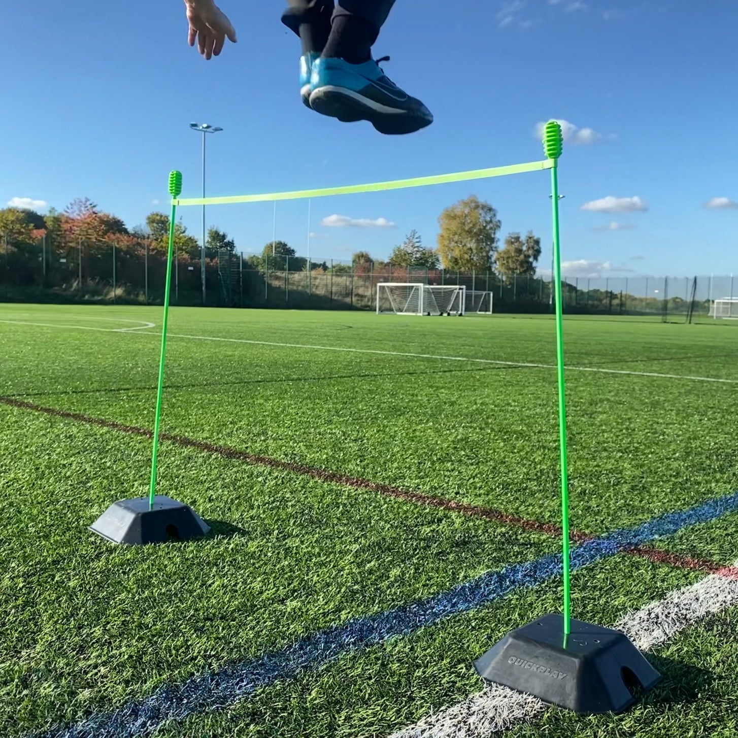 Player jumping over a green strap stretched between two agility poles standing in black Base Weights on an artificial field.