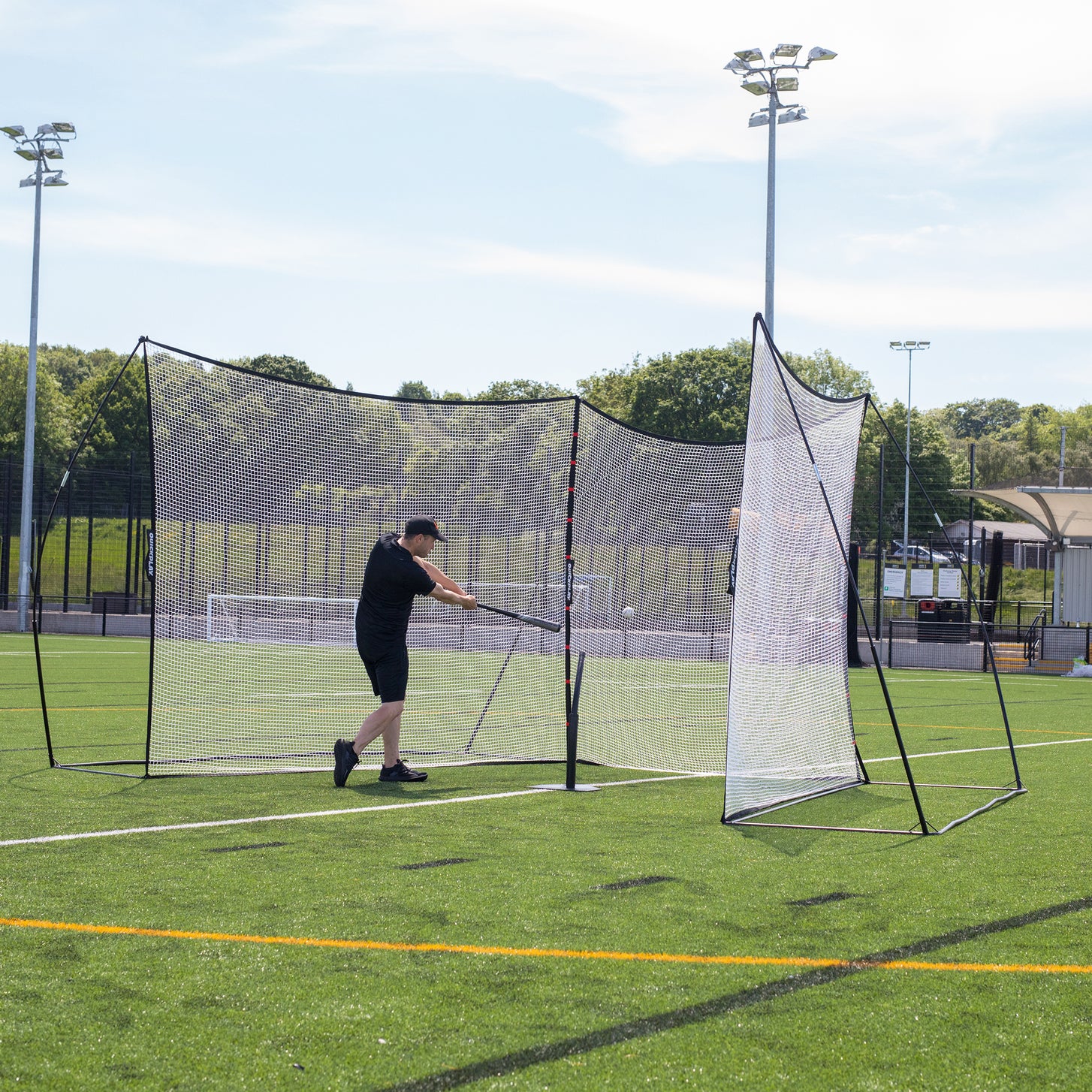 Man swinging a baseball bat in front of the MEGA NET XL Multi Sport Backstop Net 32x9' on an artificial field.