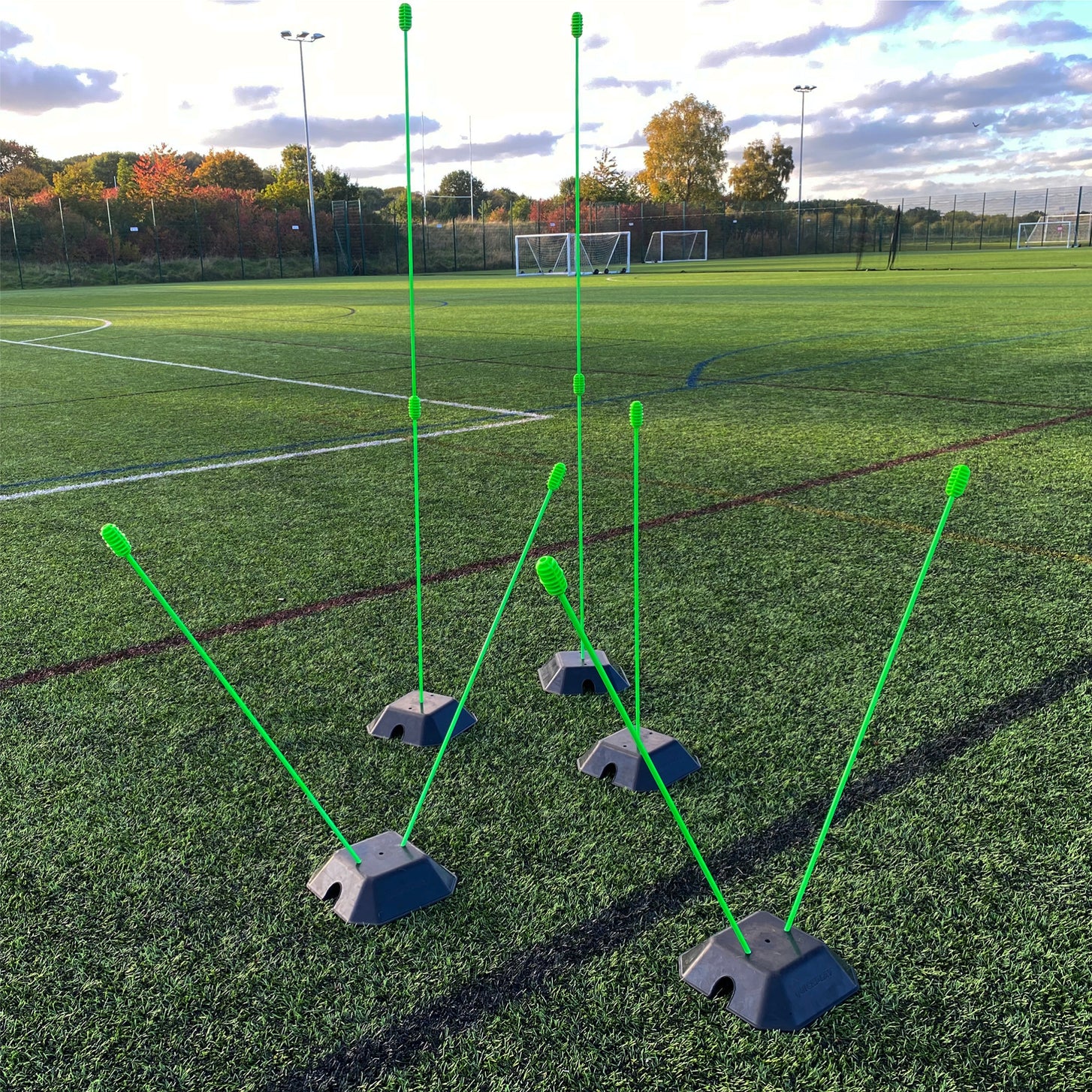 Base Weights holding green agility poles upright and at angles on an artificial field, with goals in the background.